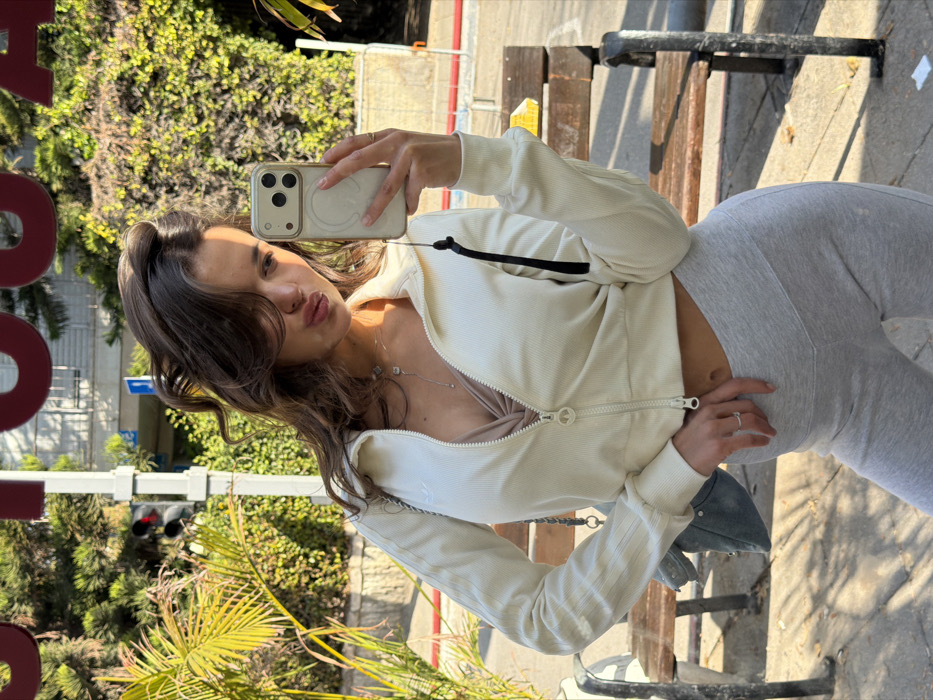 A young adult female is depicted in a full-body selfie taken in a reflective surface on an urban street in Tel-Aviv, Israel. She has wavy brown hair, partially obscuring her face, and her lips are pursed forward. She is wearing a cream-colored, ribbed, cropped zip-up jacket with three stripes on the left sleeve and an Adidas logo, partially unzipped to reveal a light brown bra or crop top. She also wears light grey leggings. A silver-colored chain necklace with a small pendant is visible, along with a silver ring on the ring finger of her left hand, which rests on her upper thigh. Her right hand holds a light-colored iPhone with a clear case and a circular magnetic accessory, from which a black lanyard hangs. A light blue studded shoulder bag hangs from her left shoulder. The background, seen as a reflection, features an outdoor urban environment with green foliage, a palm frond, two wooden benches with dark metal frames on a paved surface. A black traffic light pole with an illuminated red light and a partial blue and white street sign are visible through the vegetation on the left. Red letters, appearing as "OOJ" due to the reflection, are visible at the top center. The scene is illuminated by sunlight, casting defined shadows.