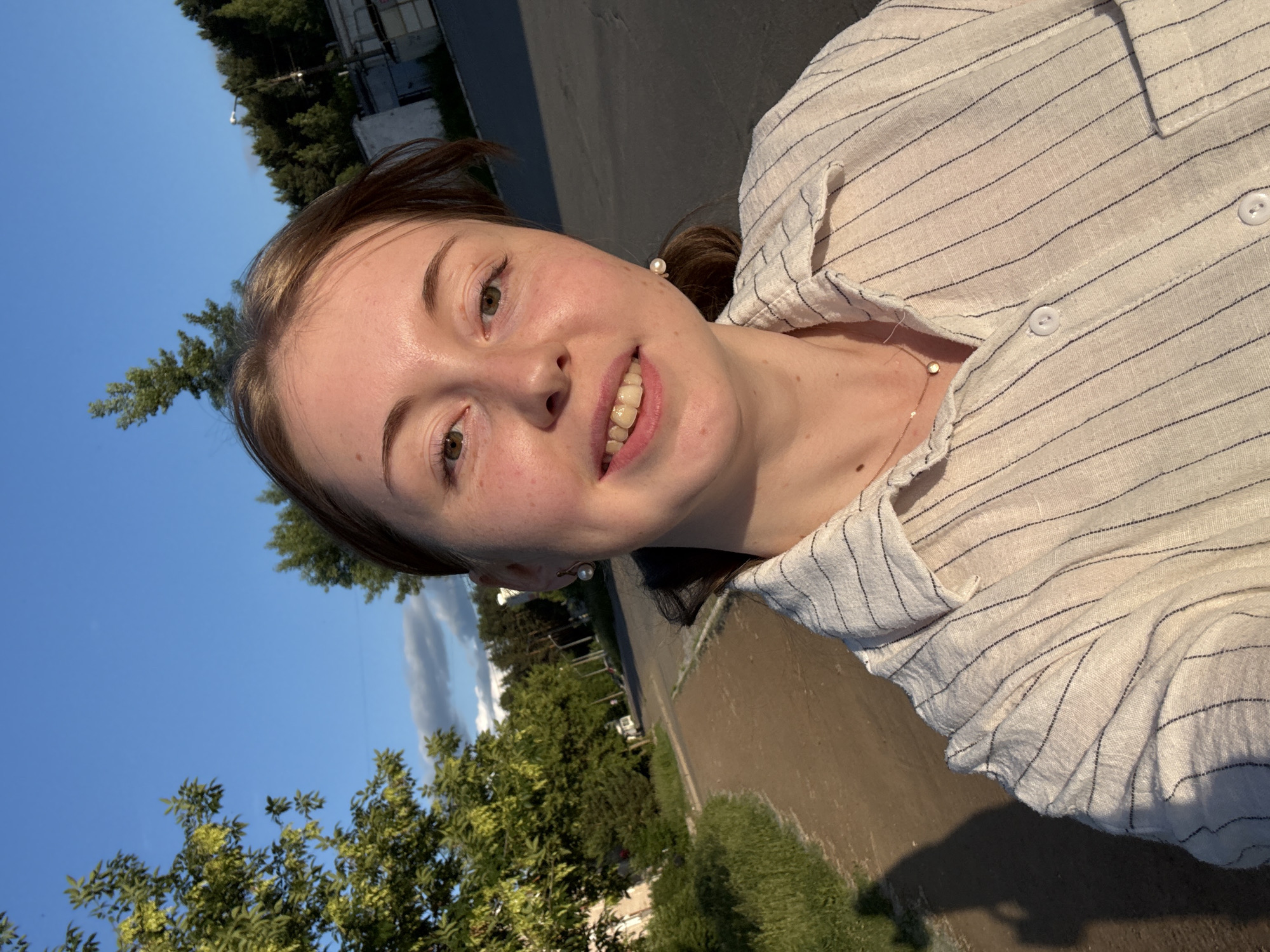 A young woman with light brown hair, fair skin, and freckles is taking a selfie outdoors during the golden hour. She is wearing a white collared shirt with thin navy blue pinstripes and a delicate gold necklace with a small circular pendant. She has a pearl earring in her visible ear. She is smiling with her mouth slightly open, revealing her teeth. The background shows a paved pathway next to a dirt path, bordered by green grass and trees. Beyond the trees, there are buildings and more trees, suggesting a suburban or semi-rural environment. The sky is clear blue with a few wispy clouds. The lighting indicates it is late afternoon or early evening.