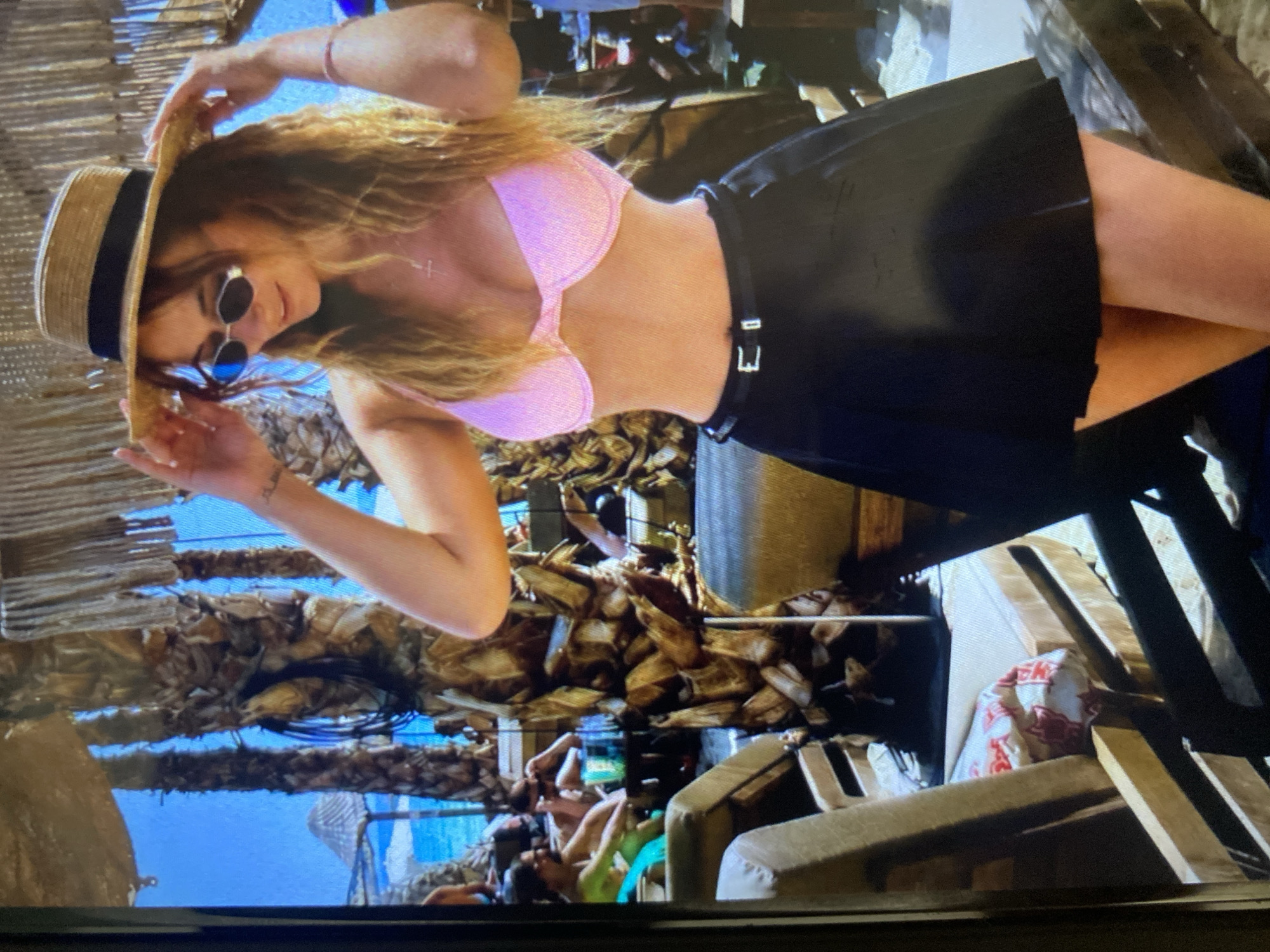 The image, rotated 90 degrees counter-clockwise, features a young woman posing in an outdoor, sunny setting. She is positioned slightly to the right, adjusting a straw hat with a dark band on her head. Her long, wavy, light brown or blonde hair flows around her shoulders. She wears a shimmering light pink bikini top and a black pleated mini skirt, fastened with a black belt that has a silver buckle, exposing her midriff. Round, dark sunglasses cover her eyes, and a small, silver-colored cross pendant hangs from a necklace around her neck. A faint tattoo is visible on her left forearm/wrist.
The background shows a leisure or resort-like environment. Numerous thatched structures, likely roofing or shading, are prominent, creating a rustic, tropical aesthetic. Beyond these, a clear, bright blue sky dominates, suggesting a warm and sunny day. In the middle distance, blurred figures of other individuals can be seen relaxing on loungers or daybeds. Various wooden elements, including chairs and structural beams, are also present. In the far right background, a white bag with indistinct red text, possibly including "CK," is partially visible. The overall scene conveys a relaxed, vacation-like atmosphere, likely at a beach or resort, within the given context of Sofia, Bulgaria.