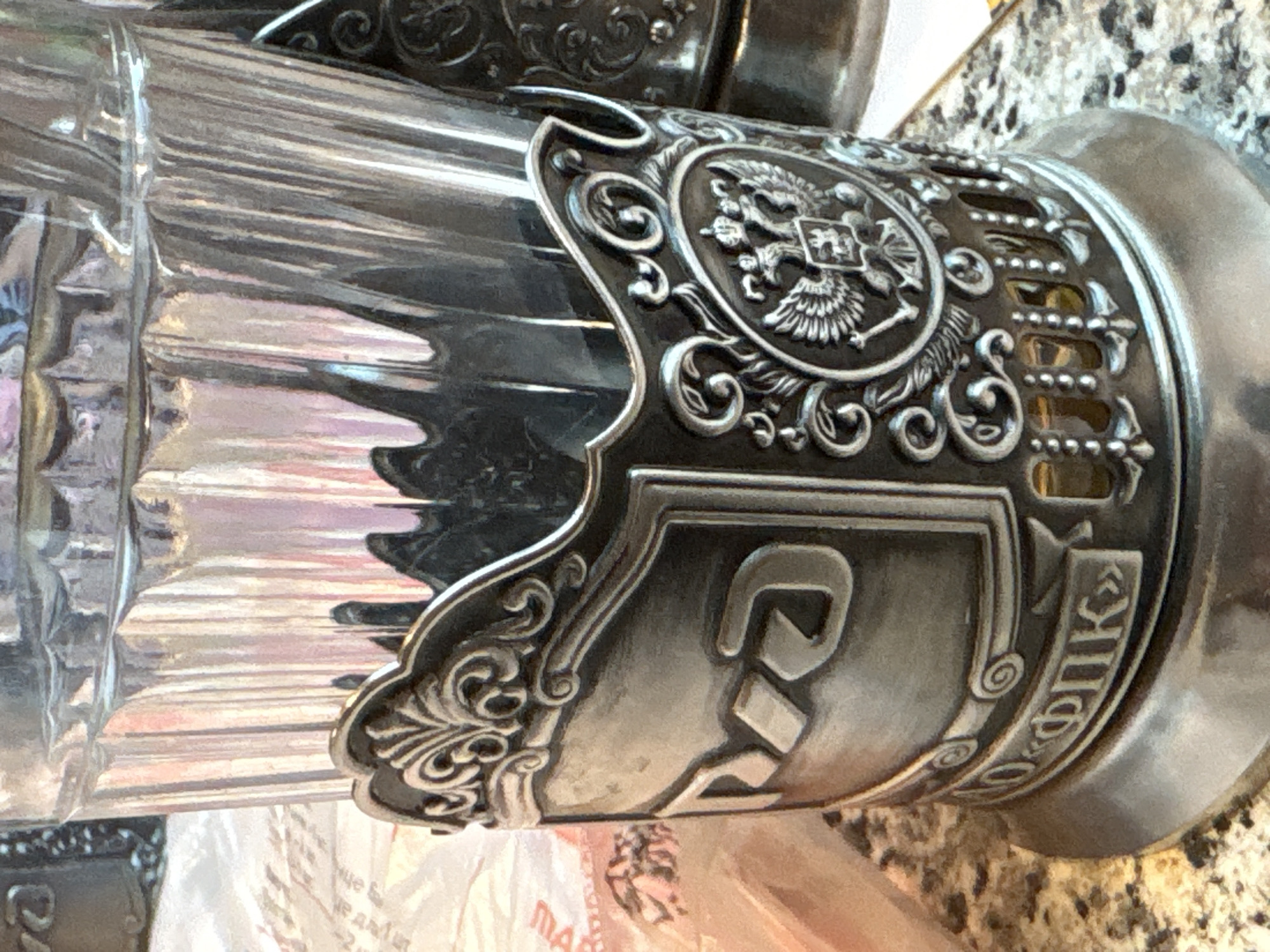 This media file is a close-up shot of a decorative metal holder for a glass. The holder is ornate, featuring embossed patterns and what appears to be a double-headed eagle emblem, suggesting a Russian or imperial design. The glass itself is made of clear, ribbed material, with reflections visible on its surface. The metalwork is detailed with scrolls and borders, and some of the lettering in a Cyrillic script is visible. The background is blurred but shows a speckled surface, possibly a countertop, and some lighter-colored material, possibly paper or fabric. The lighting is artificial, creating highlights and shadows on the metallic surface. No people are visible in the frame. The image is focused on the intricate details of the object, highlighting its craftsmanship. The Cyrillic text visible on the metal holder may provide clues to its origin or purpose, possibly indicating a brand or a specific historical reference. The overall impression is of a decorative and possibly antique item.