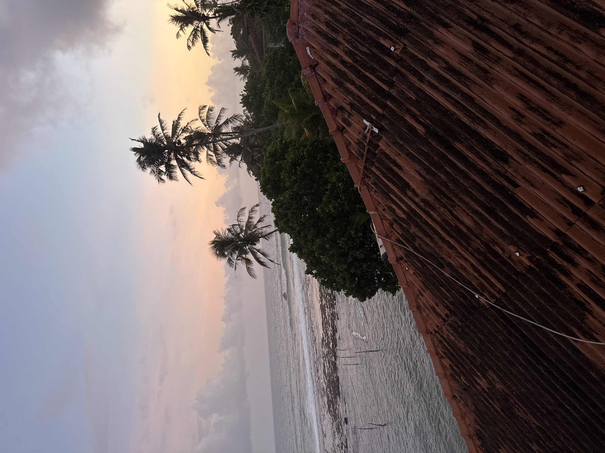 The image captures a serene coastal scene at sunset in Goviyapana, Sri Lanka. In the foreground, a weathered, reddish-brown tiled roof slopes diagonally from the lower right to the upper left of the frame, showing signs of age and some mossy growth. Beyond the roof, a lush green expanse of trees and palm trees rises, silhouetted against a soft, pastel-colored sky. The sky transitions from a pale blue at the top to hues of pink and orange near the horizon, with scattered grey clouds. The ocean stretches out to the left, its surface reflecting the muted colors of the sky. Waves gently break on a rocky shoreline, and several wooden poles are visible in the shallow water, likely used for traditional stilt fishing. The overall atmosphere is tranquil and evokes the beauty of a tropical evening.