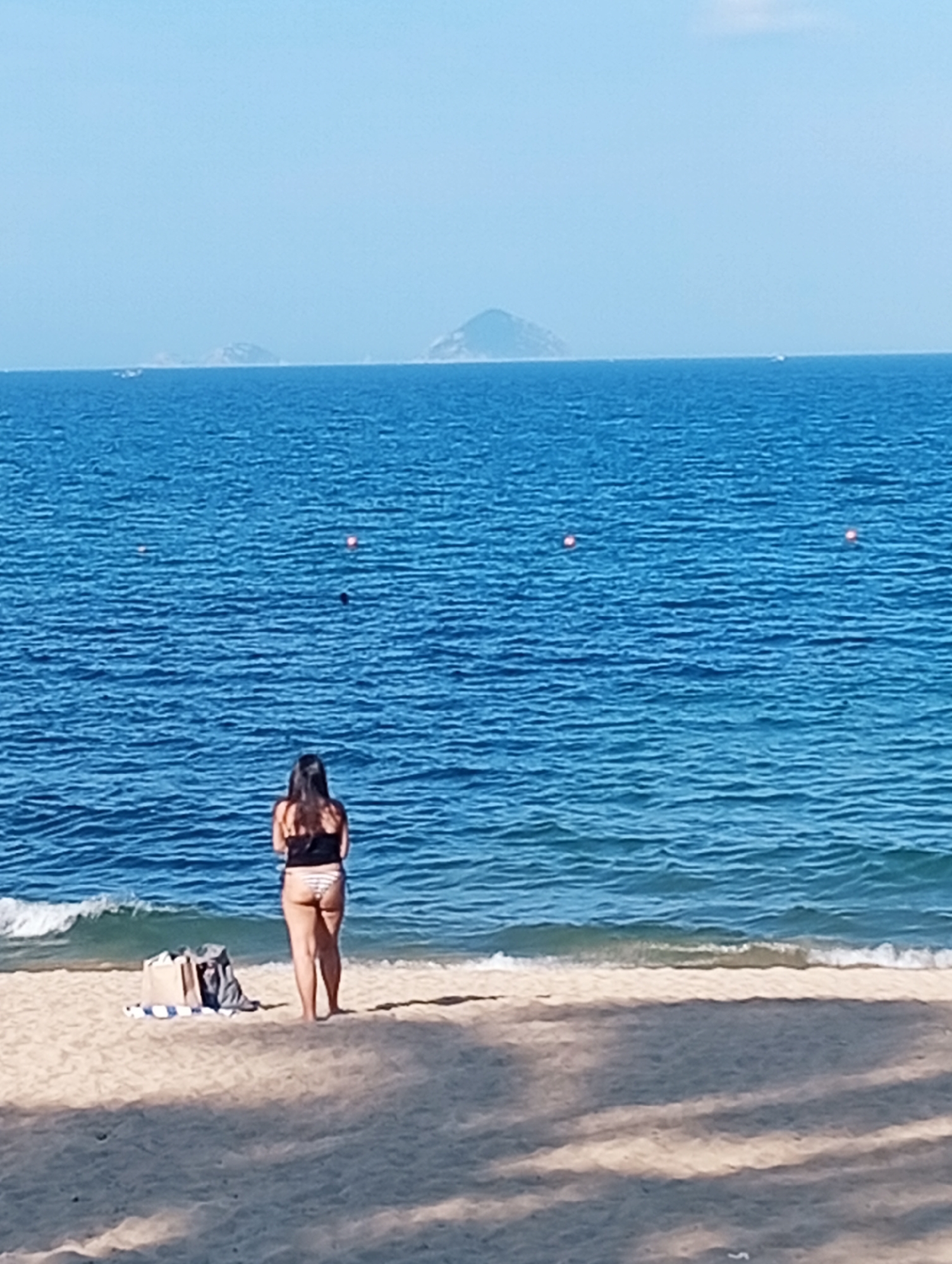 A woman stands on a sandy beach, facing the ocean. She is wearing a black tank top and striped bikini bottoms. Her back is to the camera, and her dark hair is down. In front of her, on the sand, are a couple of bags. The ocean is a deep blue with small waves breaking near the shore. There are several orange buoys visible in the water. In the distance, there are islands on the horizon. The sky is clear and blue, suggesting it is daytime with good weather. The scene is peaceful and evokes a sense of relaxation and enjoyment of the beach.