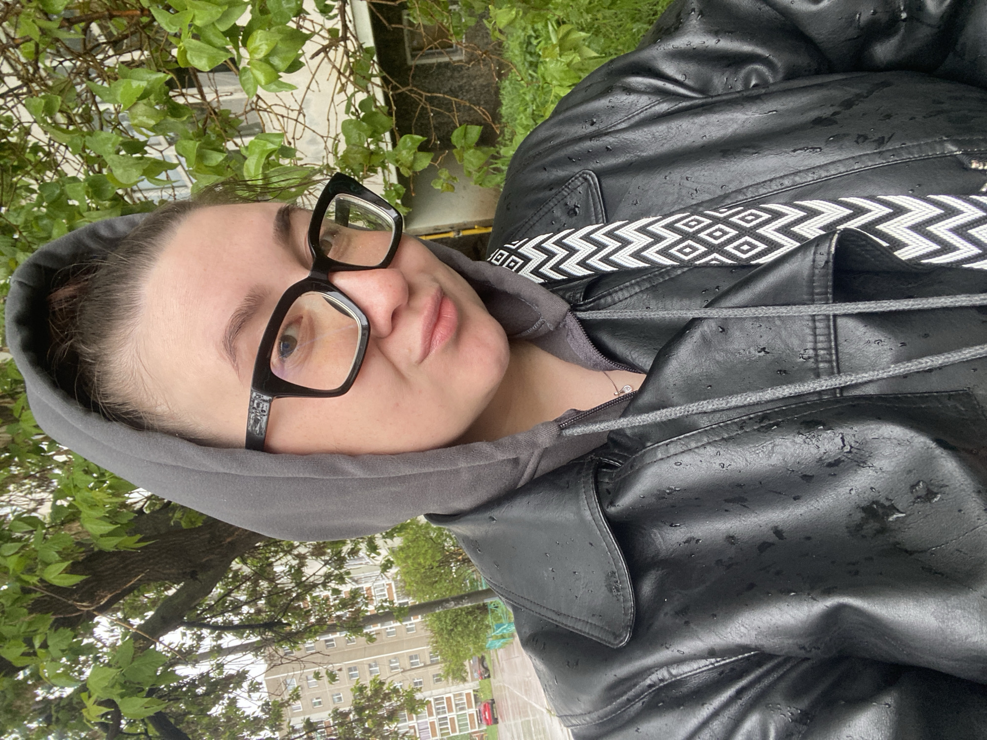 A person wearing glasses, a grey hooded sweatshirt, and a black leather jacket with a geometric-patterned strap, is outdoors, possibly in a park or green space. The location is likely in the city of Chaikovsky, Russia, as stated in the prompt. Rain is visible on the leather jacket. In the background, there are trees and buildings, suggesting a residential area within the city.