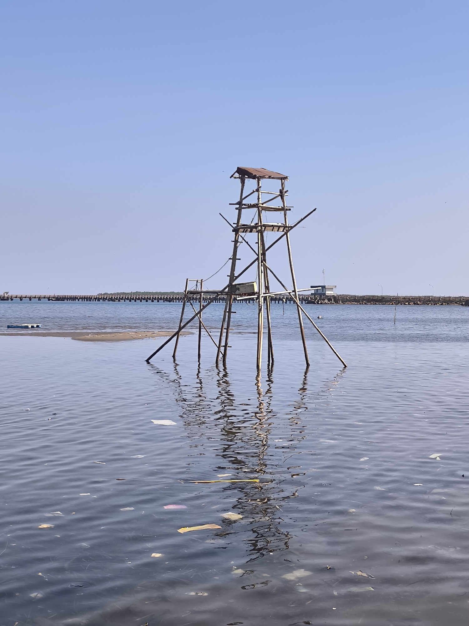 A wooden structure, resembling a lookout tower, stands in the shallow water near a sandy shoreline. The tower is made of rough wooden poles and planks, with a small, rusted metal roof on top. There are various platforms and railings constructed from wood. In the foreground, the water is calm and reflects the sky and the tower, with numerous pieces of trash, including plastic bags and wrappers, floating on the surface. In the background, a long pier stretches across the water, lined with pilings. A small white building with a dark roof sits on the shore beyond the pier. The sky is clear and blue. The location is the coastline of Barru, Indonesia.