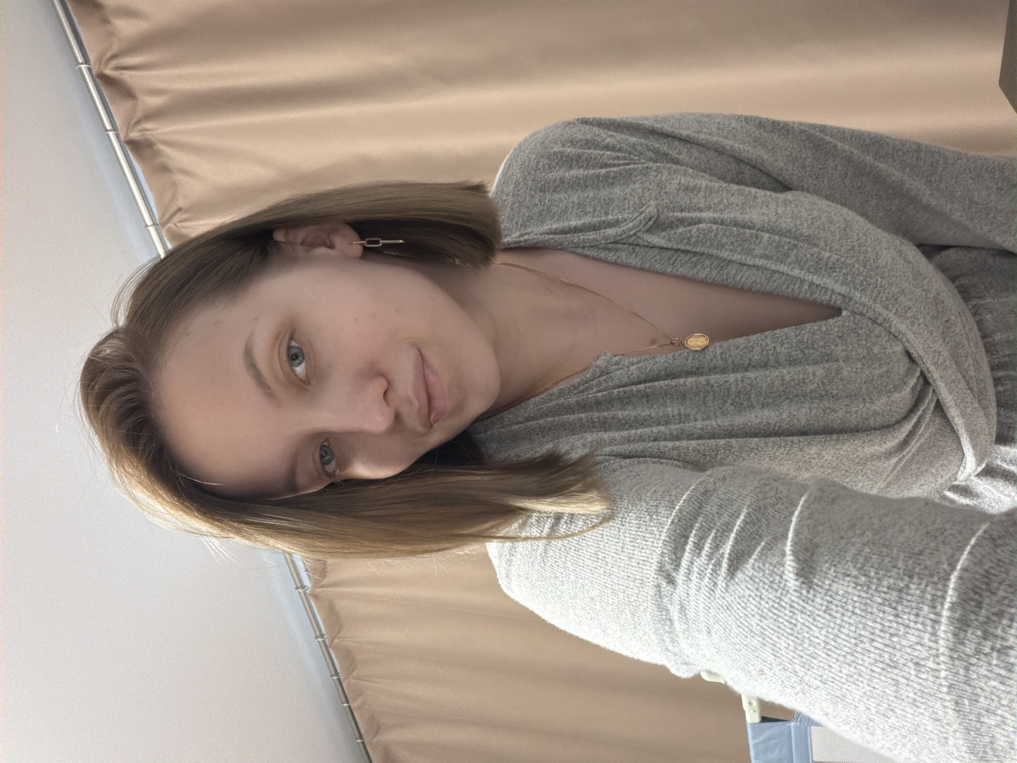 A medium shot, taken from a high angle, shows a young woman in an indoor setting. The main image is rotated approximately 90 degrees clockwise, depicting the woman lying on her side.
**Subjects:** The primary subject is a young woman with shoulder-length, light brown or dirty blonde hair and clear blue eyes. She has fair skin and a neutral to slightly soft expression, looking directly at the camera with a subtle hint of a smile. She is wearing a casual, grey, long-sleeved top made of a textured or knitted fabric, possibly a sweater or cardigan with a V-neck. A delicate gold-colored chain necklace with a small, possibly octagonal or hexagonal pendant rests on her chest. A small, minimalist gold-colored earring, shaped like a thin rectangle or bar, is visible in her right ear.
**Scene and Setting:** The background features a light beige or tan curtain or drape, hanging on a silver-colored rod, visible behind and above the woman's head. A plain white wall or ceiling fills the upper left portion of the image. A small part of a light blue or grey object, possibly a piece of furniture or container, is barely visible in the bottom right corner of the overall image.
**Activity or Event:** The woman appears to be taking a selfie, holding the camera or phone above her and looking down into the lens.
**Notable Details:** The lighting is soft and even, suggesting an indoor setting with diffused light. No text is visible in the image. This scene takes place in Reshetnikovo, Russia.