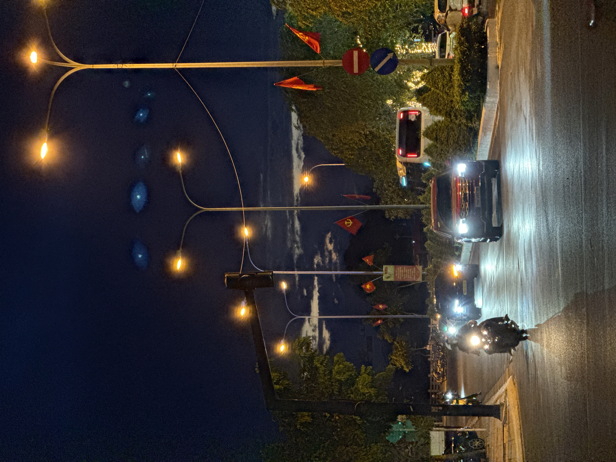 The image depicts a nighttime street scene in Nha Trang, Vietnam, likely at an intersection or along a main road. The sky is a deep, dark blue with streaks of clouds reflecting the ambient light. Several lampposts illuminate the scene with warm, orange light, casting long shadows on the wet asphalt road.
In the foreground, a motorcycle rider, wearing a helmet, is moving away from the camera, their headlight brightly illuminating the road ahead. To the right, a red SUV is approaching the viewer, its headlights also shining brightly. Behind the SUV, a white bus and other vehicles are visible, as well as decorative lights on trees lining the street.
Several Vietnamese flags are prominently displayed on poles along the roadside, some fluttering in the breeze. Traffic signs, including a red circular sign with a horizontal white bar and a blue circular sign with a white arrow, are also present. The overall impression is of a lively urban environment at night, with traffic and street decorations creating a dynamic atmosphere.