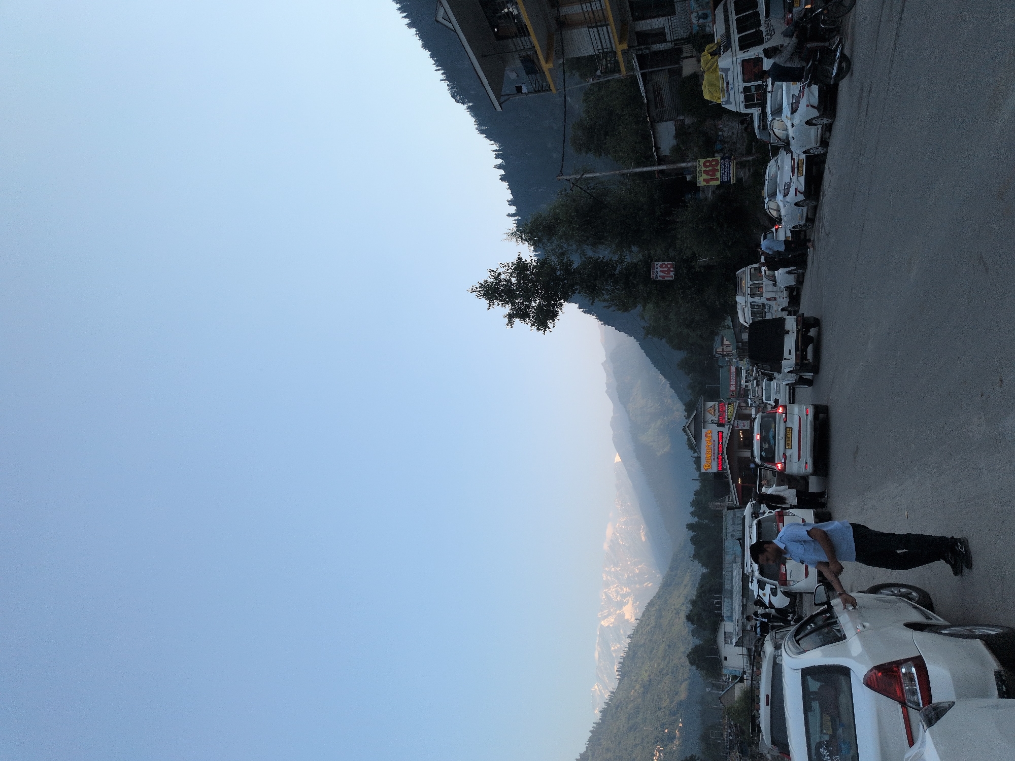 The image shows a busy street in Vashisht, India, with several cars parked and driving. The sky is clear and blue. In the background, snow-capped mountains are visible. A man in a blue shirt and black pants is standing next to a white car, appearing to open the passenger door. Another white car is parked in front of him. A line of cars, including a white van and a white SUV, is visible on the right side of the street. A white motorcycle is parked on the side of the road. A sign with the number "148" is visible on a pole. Buildings are visible on the right side of the street, with balconies and windows. Trees are present on the left and right sides of the street, and the mountains are covered with trees.