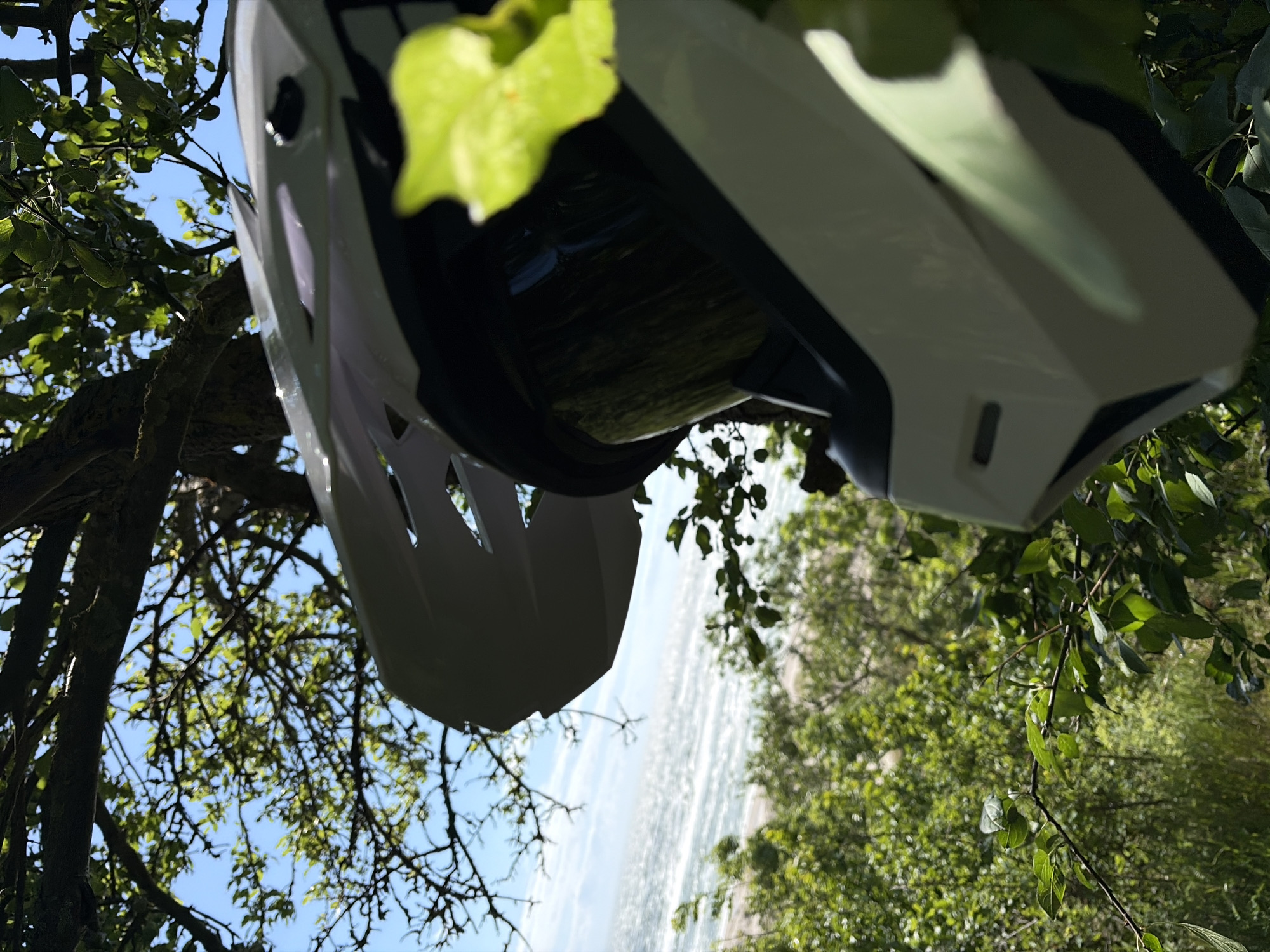 The image shows a white motocross helmet resting amongst green foliage. The helmet is positioned in the foreground, partially obscuring the background which features a bright blue sky with some scattered clouds. Beyond the foliage and sky, a body of water, possibly the sea or a large lake, is visible, with white waves suggesting it is somewhat choppy. The location is Povarovka, Russia.
