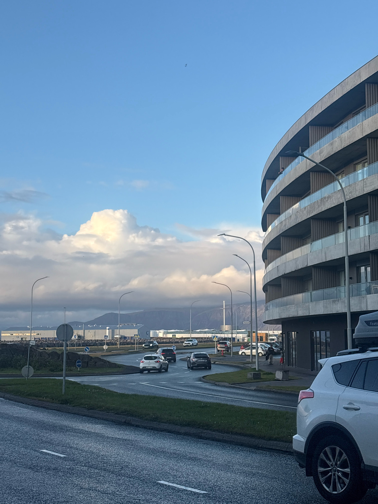 A modern, curved building with a concrete facade and multiple balconies with glass railings is situated on the right side of the image. The building appears to be residential or commercial. In the foreground, a white SUV is parked and facing away from the camera. The street in front of the building shows cars in motion, indicating traffic.
In the background, a large industrial building with a light-colored roof and multiple sections is visible. Beyond that, mountains are silhouetted against a sky filled with dramatic, puffy clouds, some illuminated by sunlight, suggesting either early morning or late afternoon. The overall environment appears to be urban or suburban, with elements of infrastructure like streetlights and road markings visible. A small bird can be seen in the sky.
This scene is located near the Grótta lighthouse in Reykjavik, Iceland, known for its unique architecture and proximity to the coast and industrial areas. The lighting and cloud formations suggest a Nordic climate.