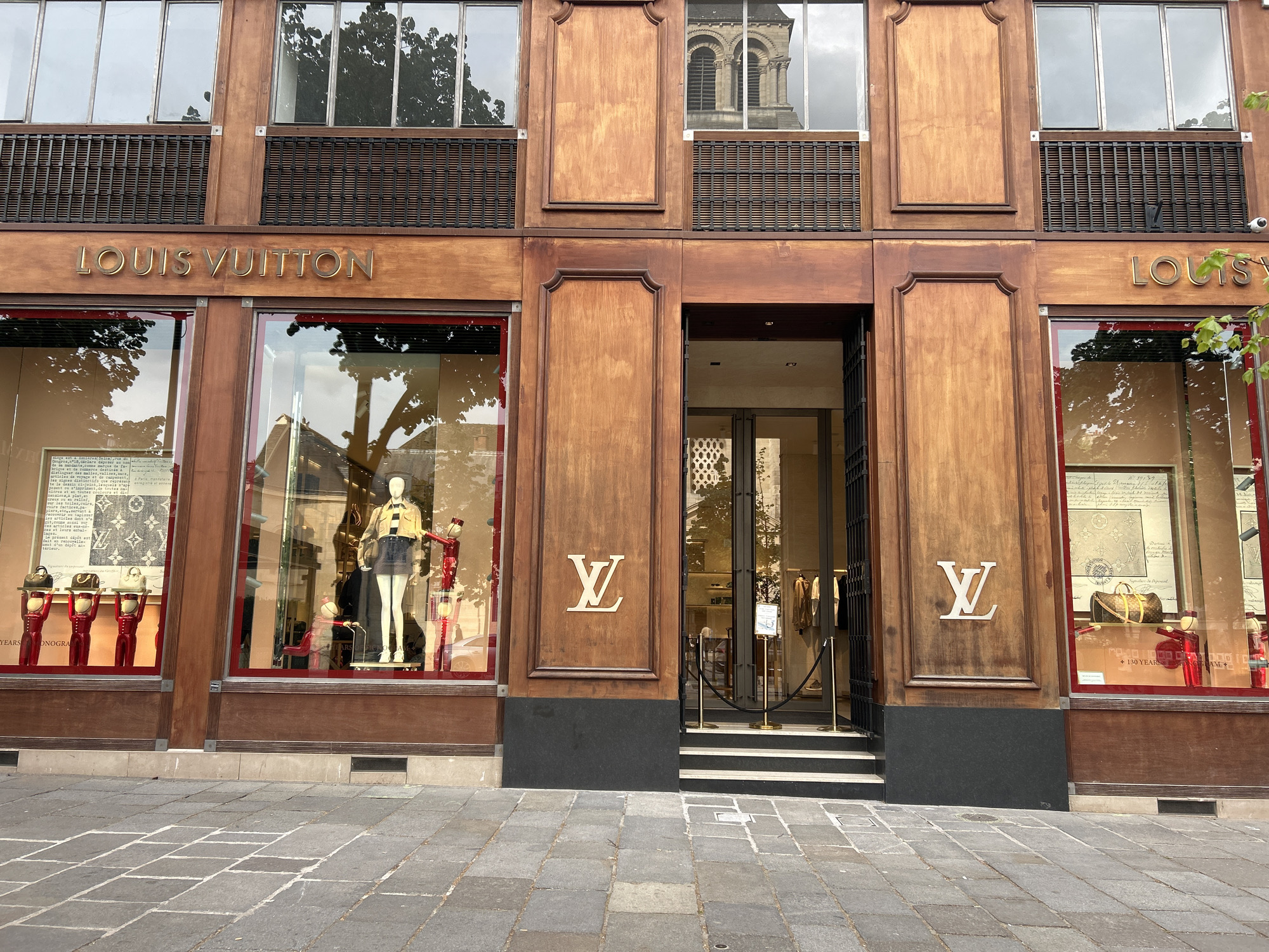 The image captures the facade of a Louis Vuitton store, likely in a city center. The building's exterior is predominantly brown wood, with large windows displaying mannequins and products. The store's name is displayed prominently above the windows. A doorway is situated centrally, with a rope barrier restricting access. The sidewalk in front is paved with gray stone blocks. The reflection in the windows suggests the presence of trees and buildings opposite the store.