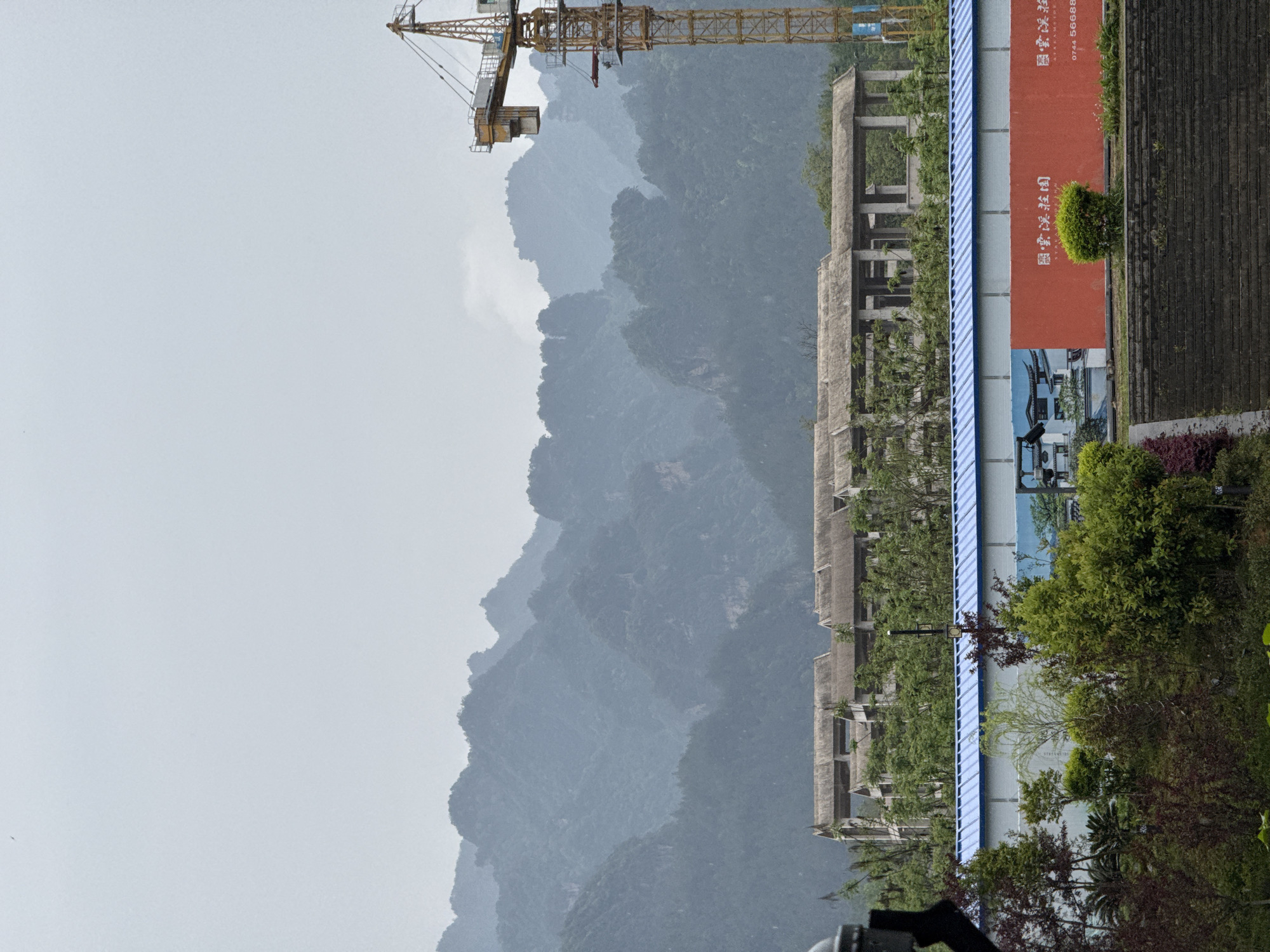 The image, oriented horizontally, presents a scene contrasting natural mountain grandeur with human development in Wulingyuan, China. In the background, majestic, misty mountains covered in dense green vegetation rise in successive layers, creating a sense of atmospheric depth under an overcast, bright grey sky.
In the mid-ground, a series of unfinished, multi-story concrete building frames stand partially obscured by trees and foliage, suggesting ongoing construction or a paused development project. A large yellow construction crane is prominently positioned in the upper left, its boom extending over the mountain range, further emphasizing the theme of development.
The foreground features a long, multi-colored advertising banner or hoarding running horizontally across the bottom. The banner has a red section with Chinese characters "国贸·深投司" (Guómào·Shēntóu Sī), likely a company or project name such as "International Trade & Shenzhen Investment Company," and a partial phone number "0744 56668". A blue section of the banner displays an illustration of a residential building and less discernible Chinese text, indicative of property advertisement. Various green trees and reddish-purple shrubs are scattered in the immediate foreground, and a dark, possibly paved area is visible on the far right.
No direct human activity is depicted. The overcast weather contributes to the soft, diffused light and the misty appearance of the mountains. The overall scene captures the intersection of natural beauty and urban expansion in a mountainous region.