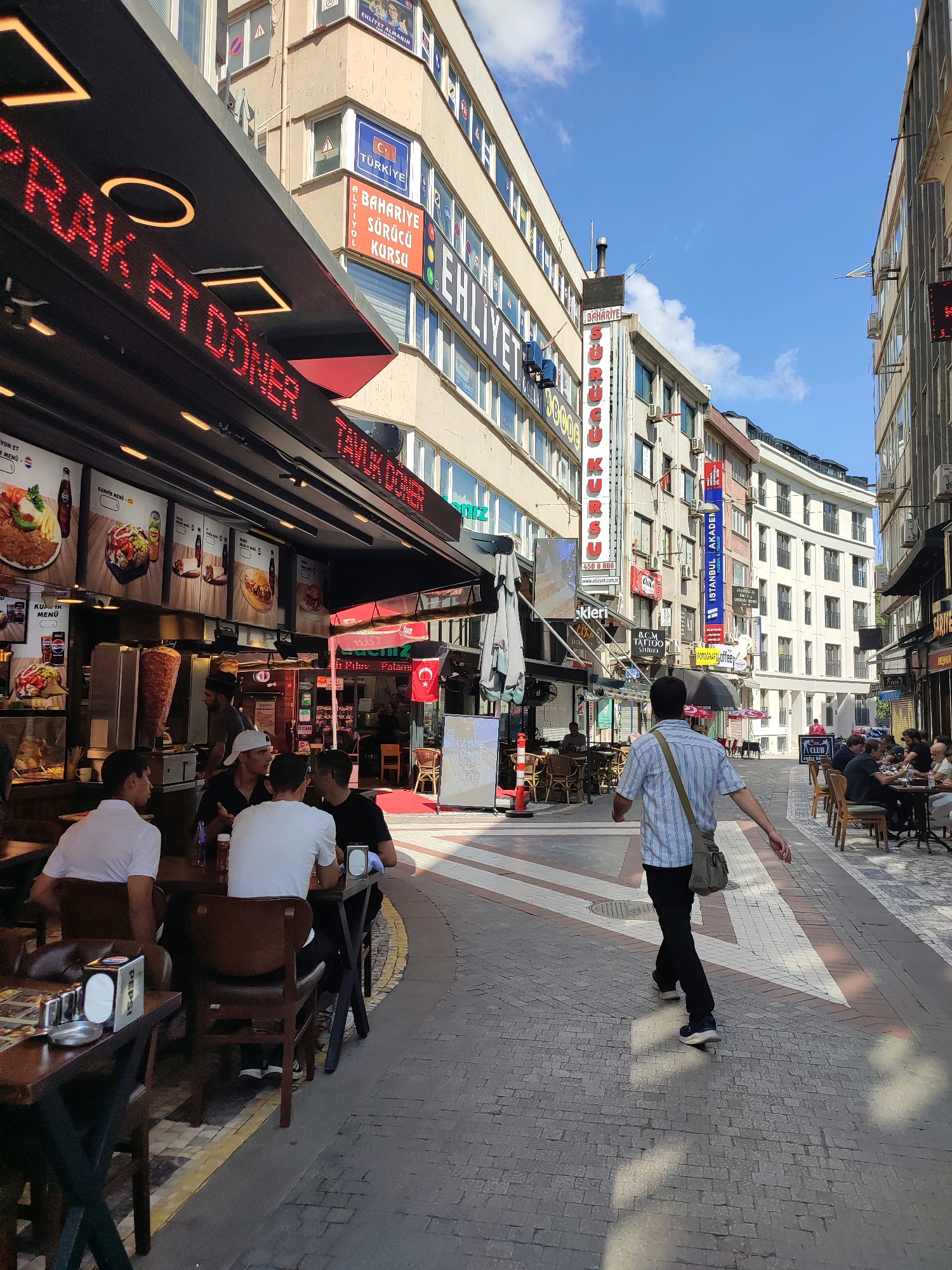 The image depicts a street scene in Kadıköy, Turkey. On the left, a restaurant with a prominent red LED sign displays "ÇIRAĞ ET DÖNER". Inside, a large vertical spit of doner meat is visible, along with food advertisements on display boards. Several patrons are seated at tables outside the restaurant. In the center and right of the image, the street continues, lined with multi-story buildings featuring various businesses and signage, including "İSTANBUL AKADEMİ" and "ACR TATTOO STUDIO". People are seated at tables outside other establishments, and a man in a striped shirt and dark pants is walking away from the camera. The ground is paved with cobblestones and decorative patterns. The sky is blue with scattered clouds, indicating daytime.
