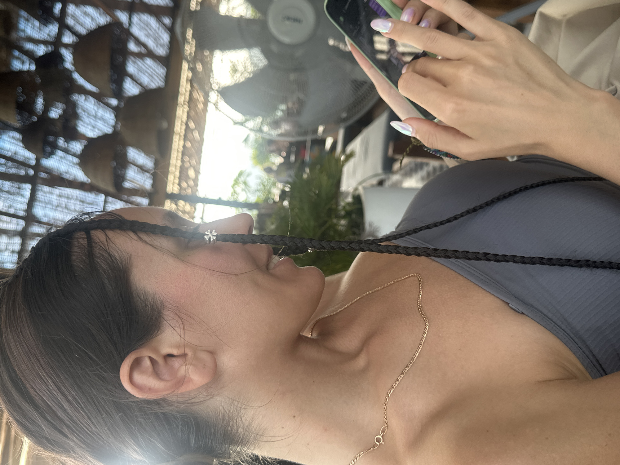 A close-up, portrait-oriented photograph, rotated 90 degrees clockwise, captures a person engrossed in their smartphone in what appears to be a casual outdoor or semi-outdoor setting in Hà Giang 1 Ward, Vietnam.
The subject is seen in profile from the chest up, with dark hair and a relaxed posture. Their hair is styled with thin, dark braids, one of which features a small, silvery, four-leaf clover-like charm near their face. A delicate golden chain necklace adorns their collarbone area, and they are wearing a grey ribbed top. Their hands, with long, iridescent, light-colored fingernails, are visible in the upper right, actively holding and interacting with a smartphone encased in a light green cover.
In the background, a large, out-of-focus electric fan with a protective grill is visible, indicating a warm climate. Above and behind the fan, a slatted wooden or bamboo structure suggests a roof or ceiling, contributing to the impression of an open-air establishment like a cafe or restaurant. Blurred green foliage is also present further in the background.
The scene is brightly lit by strong daylight, creating prominent highlights and some lens flare, particularly around the subject's ear and hair, suggesting a sunny day. No discernible text is visible in the image.