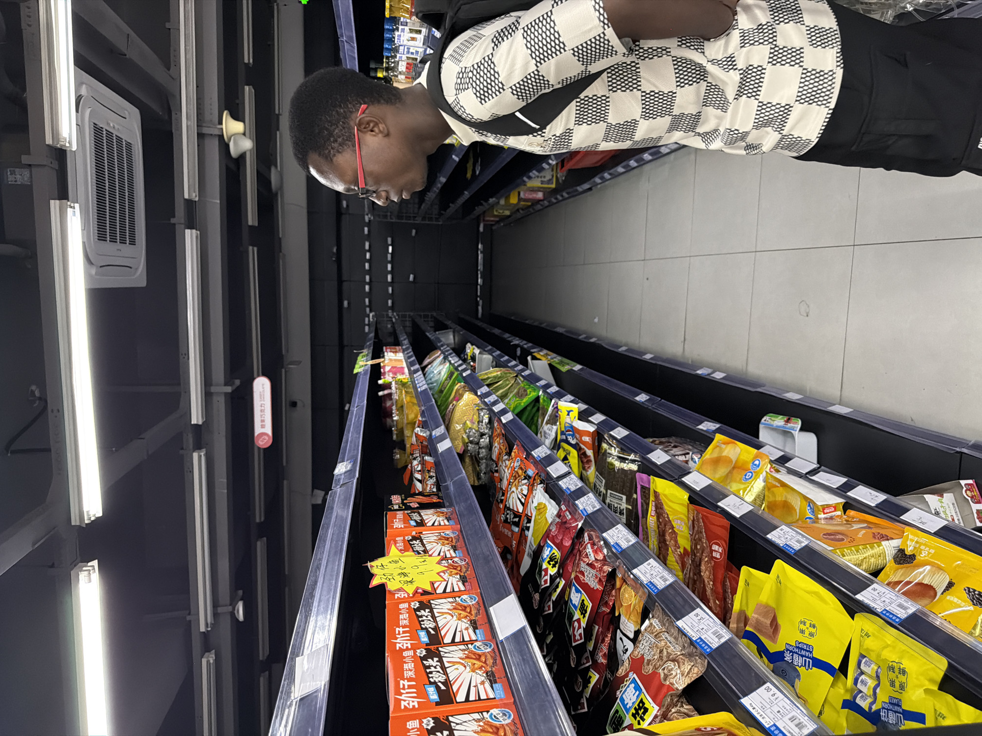 A young Black man with glasses and a checkered shirt stands in a grocery store aisle, facing shelves stocked with snacks. He is looking to his left at the products on the shelves. The aisle is well-lit by fluorescent lights, and the shelves are dark and modern. The man is wearing a black backpack and black cargo pants. The products on the shelves appear to be packaged snacks, including bags of chips and boxes of cookies. The flooring is tiled. The scene is likely in a supermarket in Jiangjin District, Chongqing, China.