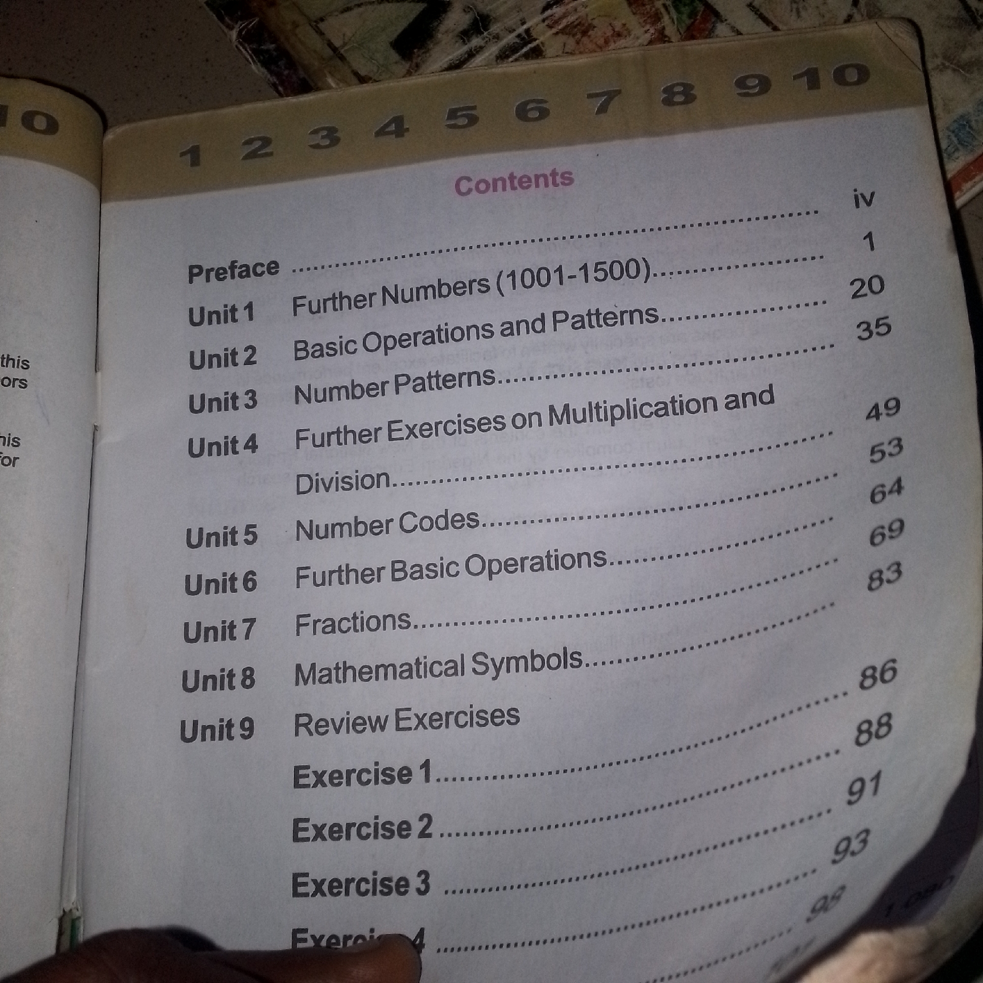 The image shows a table of contents from a book. The book appears to be a mathematics textbook, as indicated by the titles of the units such as "Further Numbers," "Basic Operations," "Fractions," and "Mathematical Symbols." The table of contents lists units 1 through 9, along with page numbers. The book is open to a page with the heading "Contents" printed in a pinkish-purple font. The page is bordered by a thick, light brown strip at the top, which has numbers 1 through 10 printed on it. The lighting in the image suggests it was taken indoors, possibly in a dimly lit room, with the flash from the camera illuminating the page. The book is lying flat, and a portion of a hand is visible at the bottom of the image, suggesting someone is holding or browsing the book. The background is a textured surface, likely a table or floor. There is no information in the image to suggest the location is in Jauro Bamvo, Nigeria.