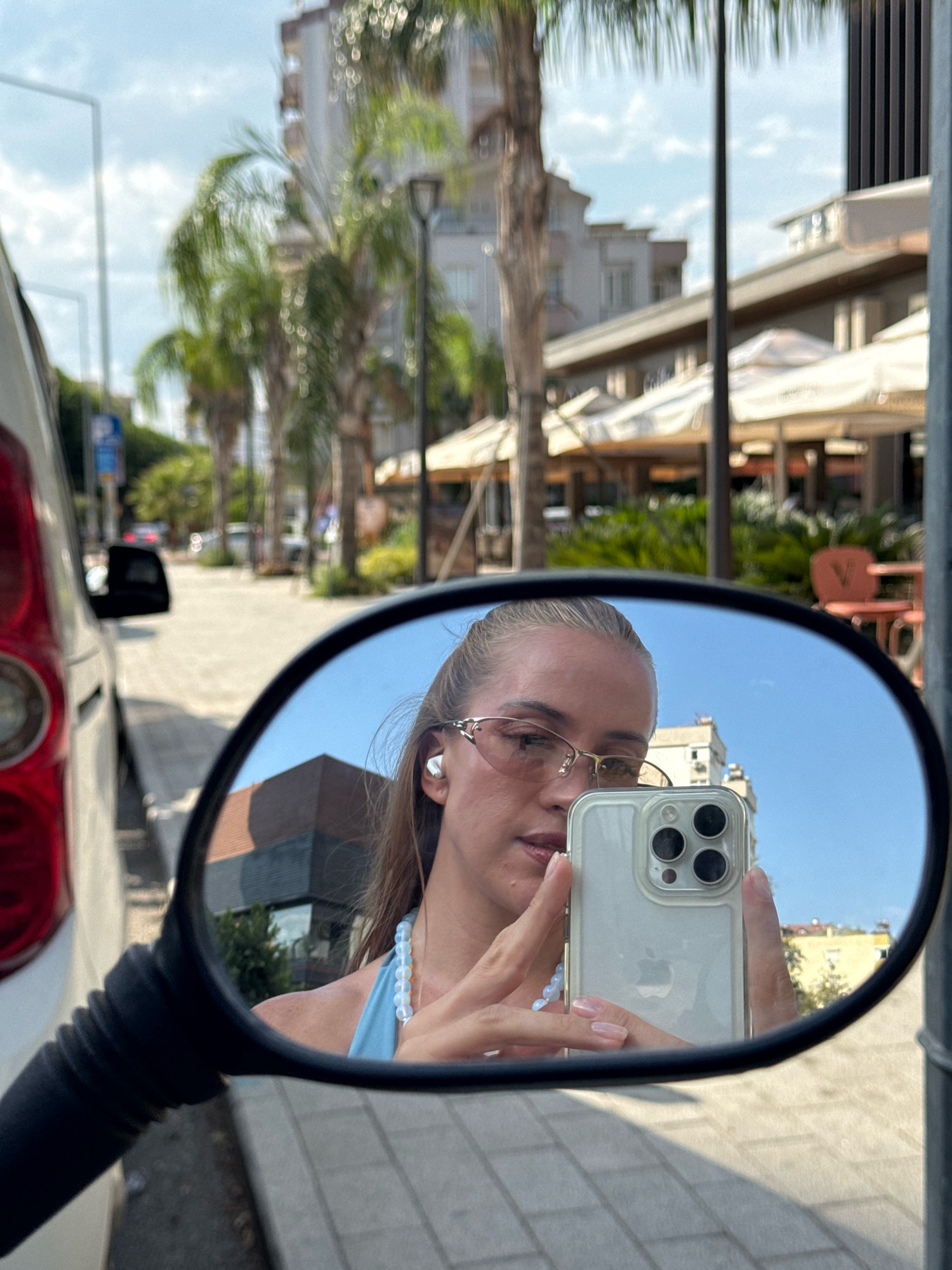 A young woman with blonde hair pulled back is taking a selfie in the mirror of a vehicle. She is wearing glasses and earbuds, and a blue top with a beaded necklace. Her phone, an iPhone, is held up to take the photo. The mirror reflects the woman, her phone, and the surrounding environment.
The background shows a city street with palm trees, buildings, and umbrellas over outdoor seating areas, consistent with a location in Antalya, Turkey. The pavement is paved with interlocking stones. The sky is blue with scattered clouds. The vehicle's rear red taillight is visible on the left side of the image.