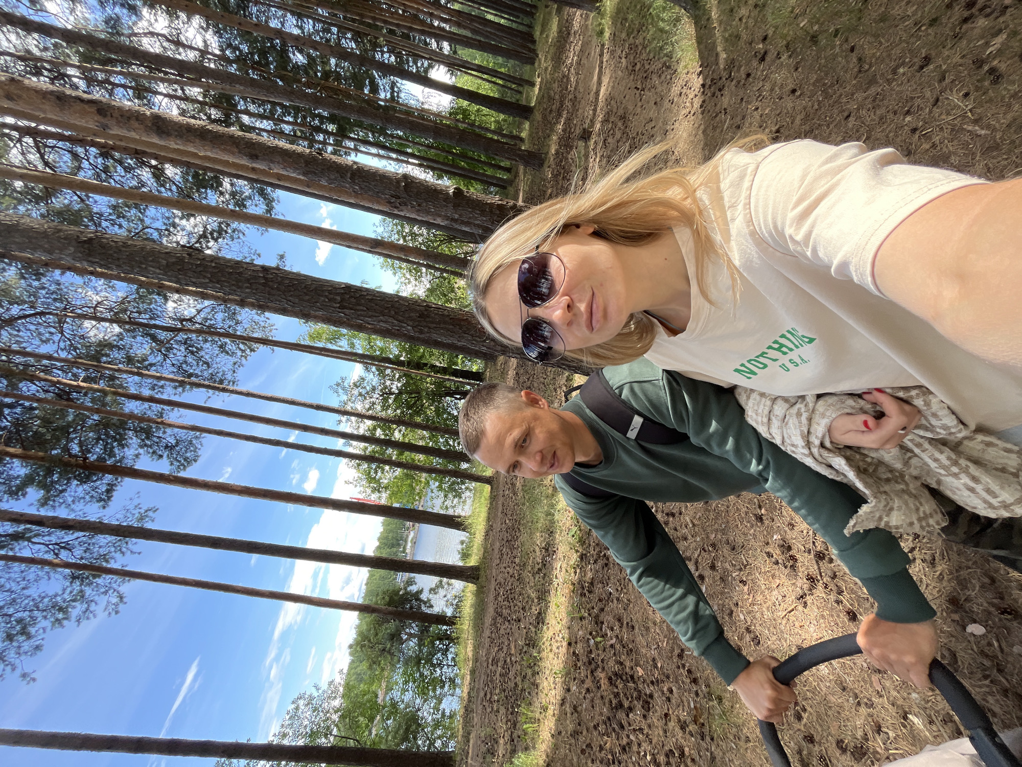 This media file is a selfie taken by two people in a forest near a body of water. A woman with blonde hair and sunglasses is in the foreground, wearing a light-colored t-shirt with green text that reads "NOTHING U.S.A." A man in a green long-sleeved shirt is in the background, pushing a stroller, indicated by his hands gripping the stroller handle. The scene is set in a pine forest with tall trees and a path covered in pinecones. Beyond the trees, a lake or river is visible, with a few boats and a structure on the water. The sky is blue with scattered white clouds, suggesting it is daytime and the weather is pleasant. The overall mood appears relaxed and outdoorsy, with the couple enjoying a walk. The presence of the stroller implies they may be with a child. The location context is Chaikovsky, Russia.