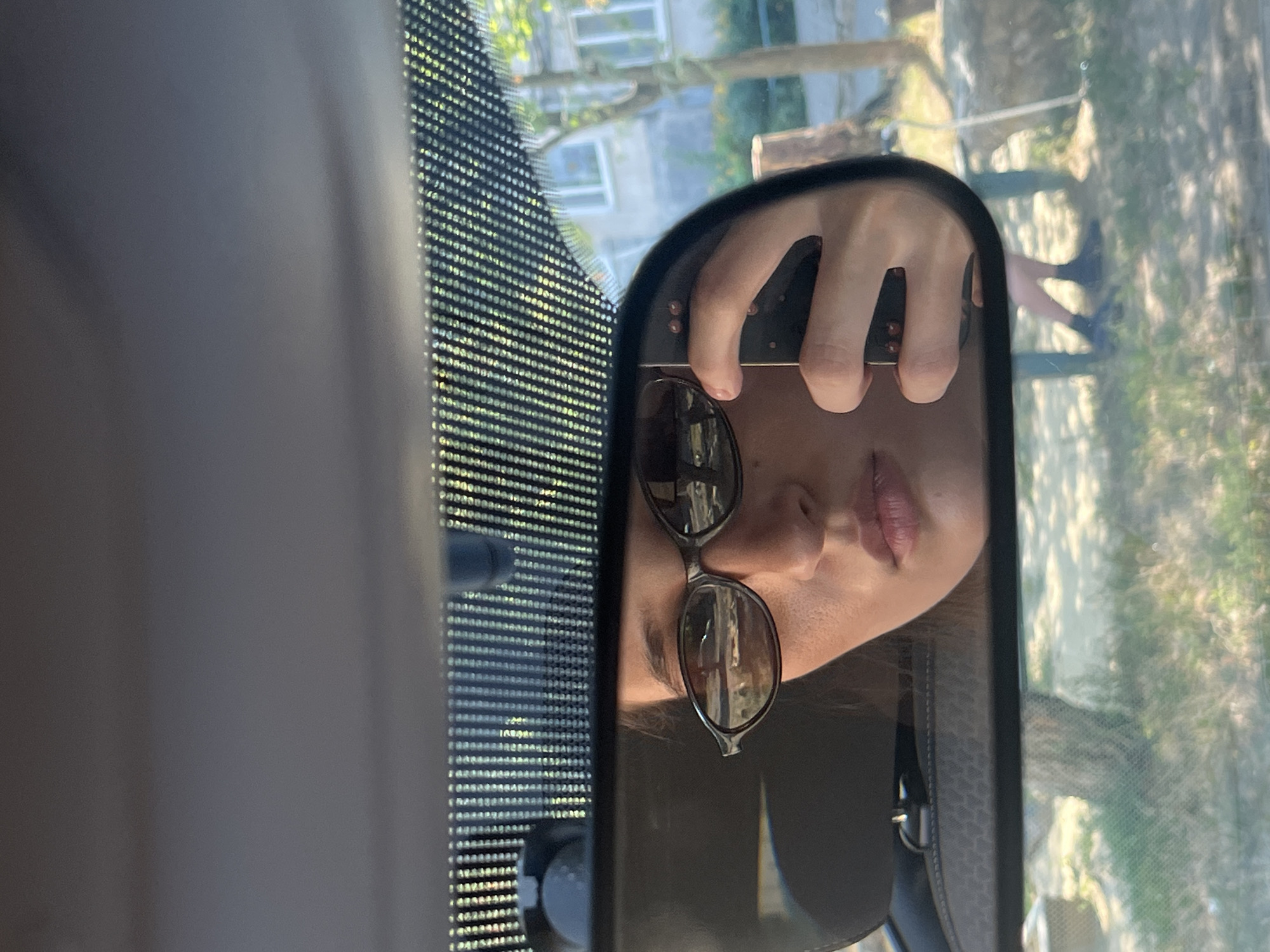 A person's reflection is visible in a car's rearview mirror. The person is wearing sunglasses and holding a phone to take a picture. The reflection shows part of the person's face, their hand, and the phone. Outside the car, a tree and a building are visible. The image appears to have been taken during the daytime.