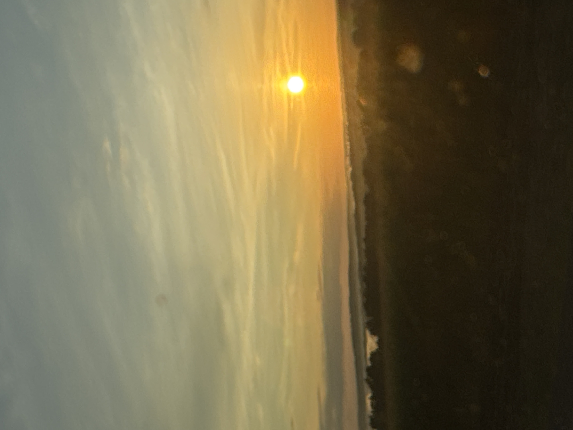 A hazy sunset over a rural landscape. The sun, a bright orb, is positioned in the upper right quadrant of the image, casting a warm orange glow across the sky. Wispy clouds stretch horizontally across the horizon, transitioning from a pale blue at the top to a soft yellow and orange near the sun. Below the sky, layers of misty hills or low mountains are visible, silhouetted against the light. The foreground is a dark, out-of-focus field, with some indistinct circular light reflections, likely lens flare, scattered across it. The overall impression is one of a serene, possibly early morning or late evening, scene in a natural setting, with no human presence or man-made structures clearly identifiable. The location is stated to be Uksunny, Russia.