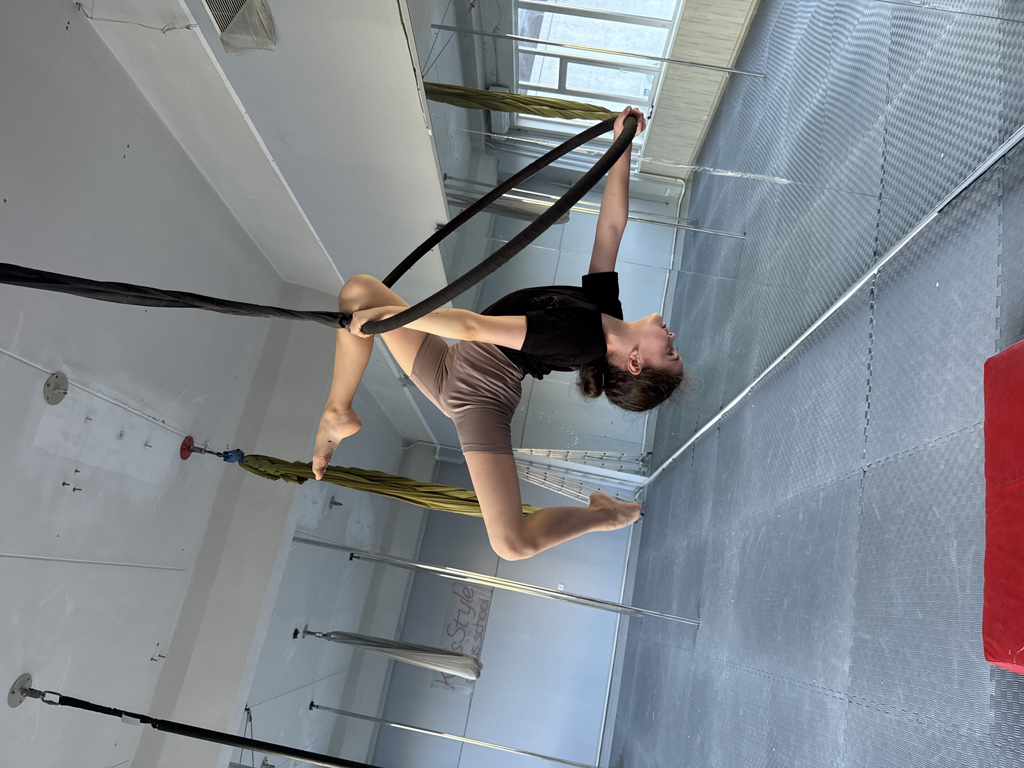 A young woman is performing aerial acrobatics in a dance studio. She is suspended by black aerial silks, with one leg extended upwards and the other bent. Her arms are wrapped around a hoop, and her body is arched. She is wearing a black t-shirt and light brown shorts.
The studio has a high ceiling with exposed beams and ventilation systems. The walls are white, and the floor is covered with grey padded mats. There are several aerial silks and hoops hanging from the ceiling, as well as some metal poles. Large windows in the background reveal a cloudy sky, suggesting it is daytime.
Visible text includes "KIDS STYLE school" on one of the poles, indicating the location is a dance school. The overall mood is one of athleticism and grace.