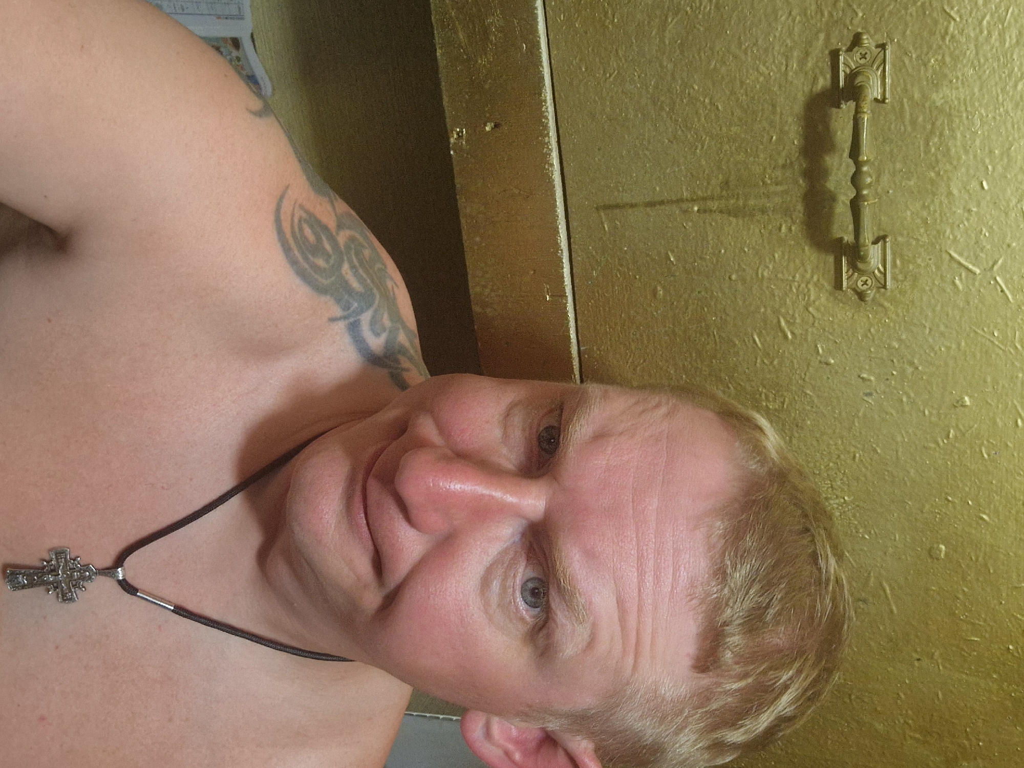 A shirtless man with blond hair and blue eyes poses for a selfie in front of a textured, golden wall. He is wearing a dark cord necklace with a silver cross pendant. A tribal tattoo is visible on his left shoulder. The background shows a brass handle and a shelf. The man appears to be in a room with a unique, possibly metallic, finish. The location is specified as Svetogorsk, Russia.
