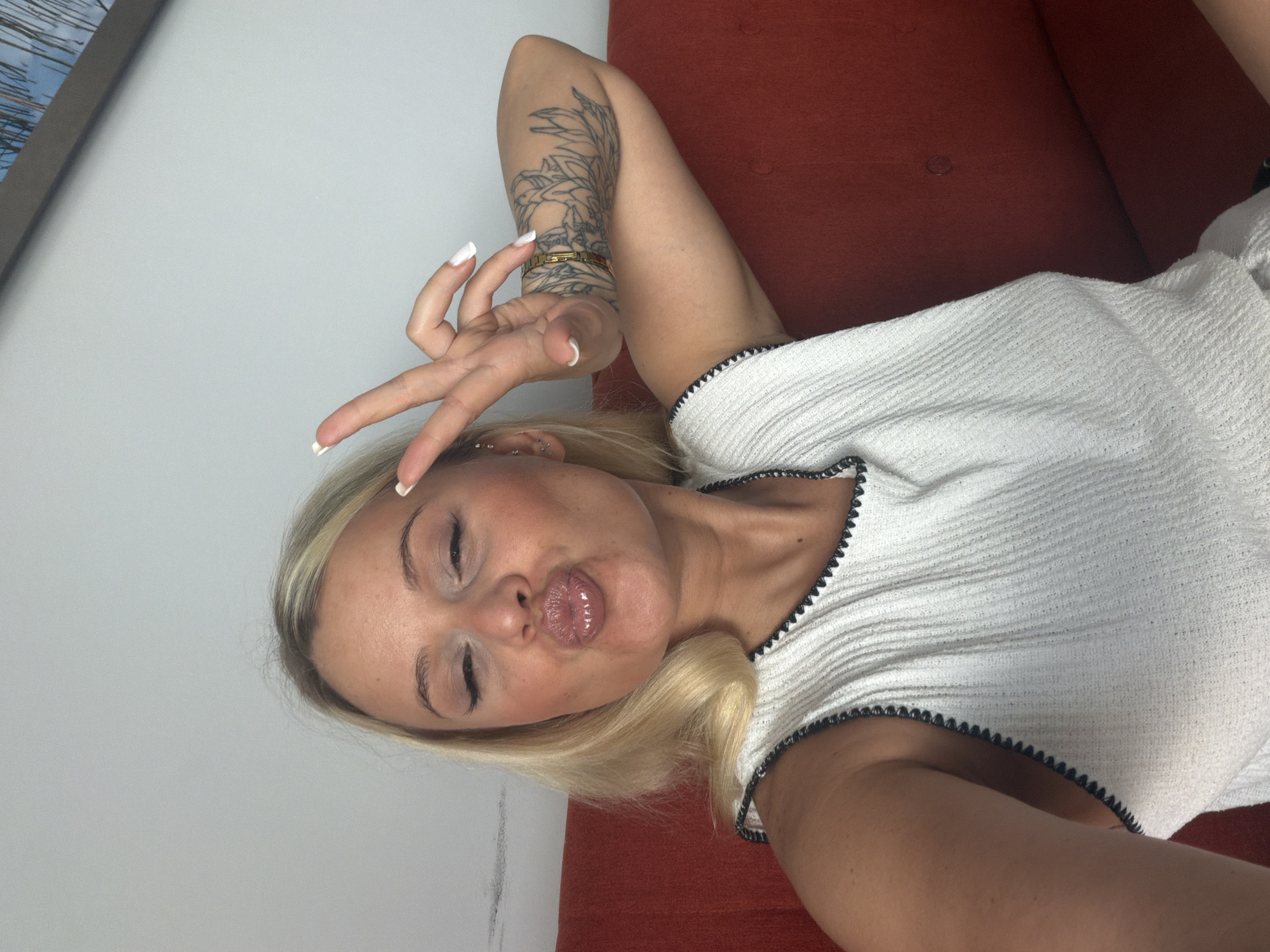 A blonde woman with a tattoo on her right arm is seated on a red couch and looking at the camera. She is wearing a white top with black trim. She is making a peace sign with her left hand and puckering her lips. The background is a plain wall. There is no information about the location in the city of Sarapul, Russia.