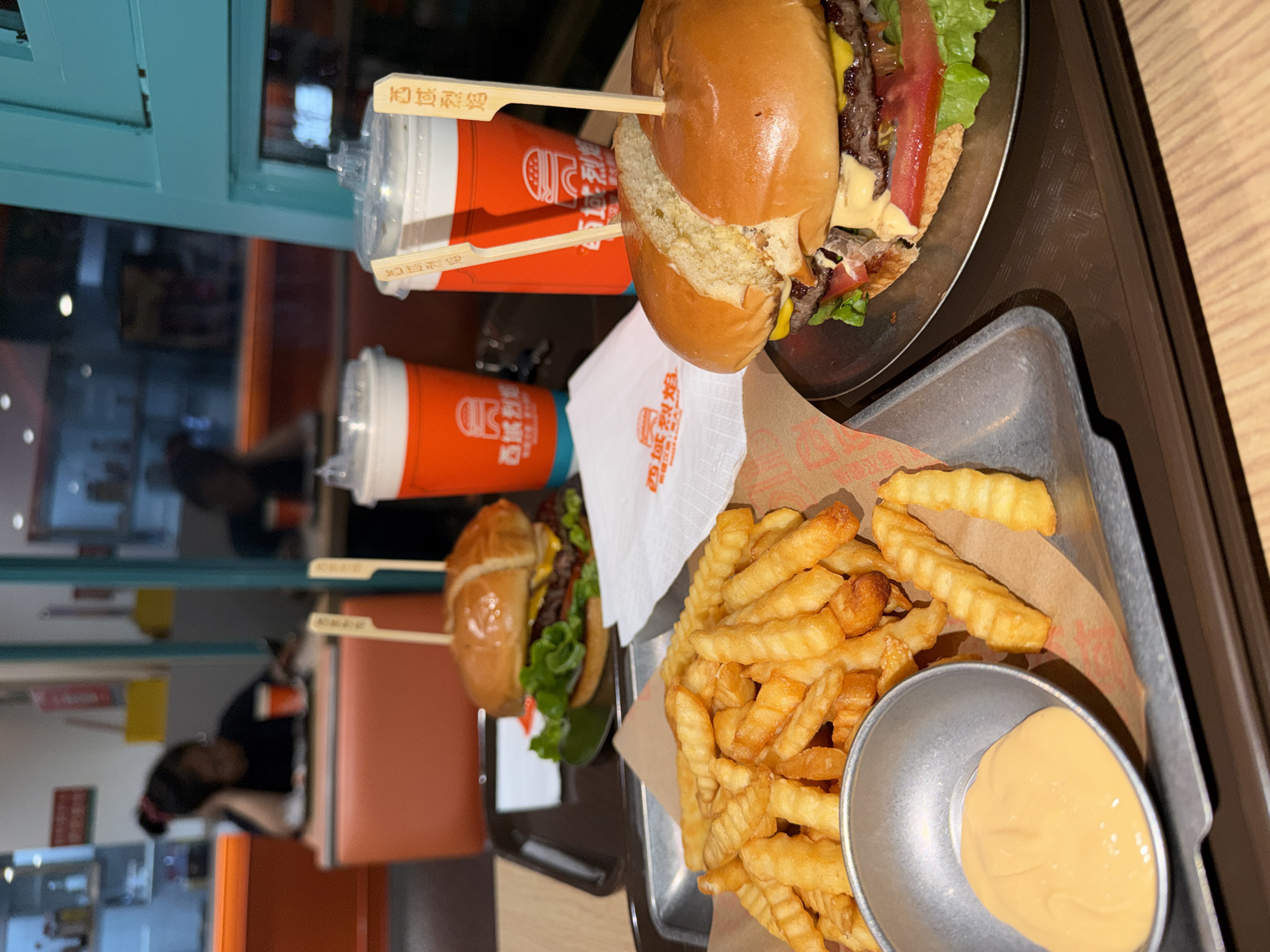 The image depicts a meal at a restaurant, likely in Chongqing High-tech Zone, China. The meal consists of two burgers on separate metal plates, a serving of crinkle-cut fries with a dipping sauce, and two orange beverage cups with chopsticks as stirrers. The food is arranged on a large brown tray, possibly made of plastic, with a metal serving tray underneath the fries and dipping sauce. A white napkin with red text is partially visible between the burgers and fries. The scene is indoors, with a window or reflective surface showing a partial view of the interior, including a seated person with their back to the camera. A red booth seat is partially visible in the background.