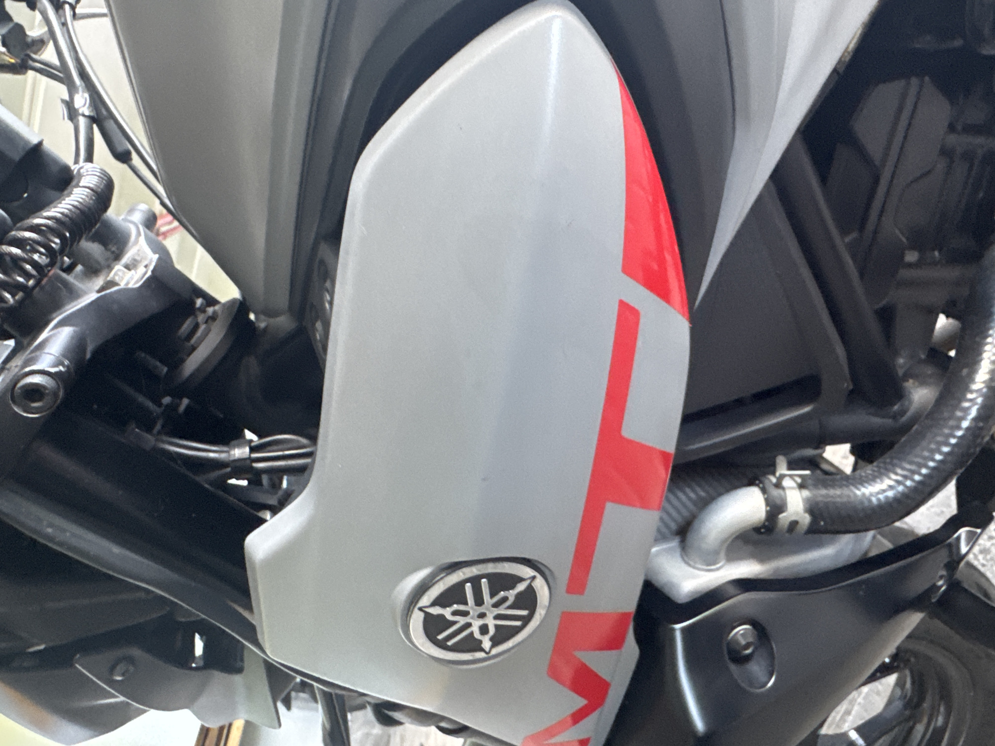 The image shows a close-up of a Yamaha motorcycle, focusing on the side fairing. The fairing is gray with a red "MT" graphic. A Yamaha logo is visible on the lower left corner of the fairing. Various mechanical components of the motorcycle are visible, including black tubes, cables, and a shock absorber. The background is blurred, suggesting a shallow depth of field. There is no information in the image to associate it with the city of North Kuta, Indonesia.