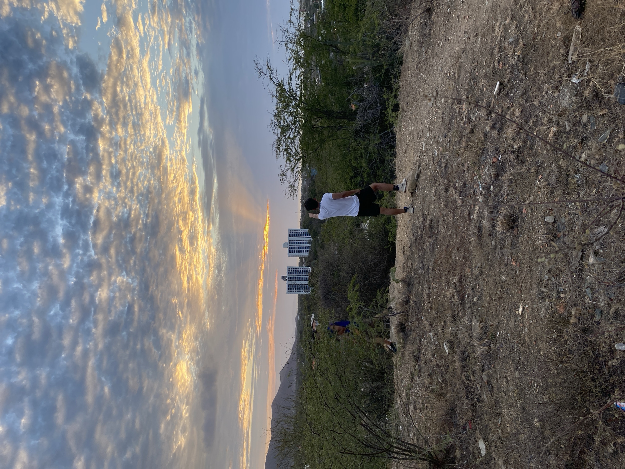 A landscape oriented photo shows two men standing on a dirt cliffside, with three tall buildings, trees, and clouds in the background. The sky is light blue with white clouds tinged with yellow, suggesting it is dusk or dawn. The ground is a dry, brown color, with some littering of white objects that could be trash. The three buildings are tall, white, and have a rectangular shape. The man closest to the buildings is wearing a blue shirt and dark shorts, but his face is indiscernible. The man closest to the top of the photo is wearing a white shirt and dark shorts; he appears to be stretching his arms out. The photo was likely taken on the Cerro Maria Guevara trail, facing the ocean, with the buildings in Juan Griego, Venezuela.