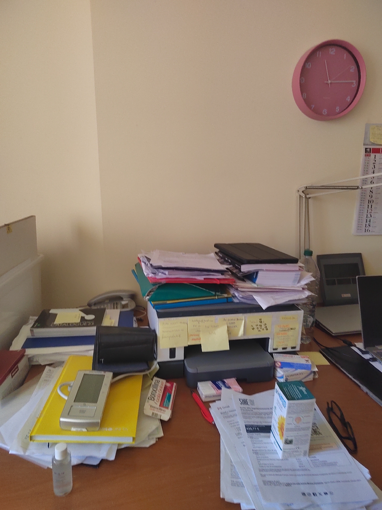 The image depicts a cluttered desk in what appears to be an office or study environment, likely located in Porto Recanati, Italy, given the context provided. The scene is dominated by stacks of papers, books, and electronic devices. A pink analog clock with black hands is mounted on a cream-colored wall in the upper right quadrant of the image. Below the clock, a calendar grid is partially visible.
On the desk, a printer is centrally positioned, with various documents and sticky notes attached to it. To the left of the printer, a stack of books and binders is visible, including a dark-colored one with the word "CATALOGUE" visible. A silver electronic device with a screen and buttons rests on a bright yellow notebook. Next to this, a small bottle of clear liquid and a pack of cigarettes are present. To the right of the printer, a laptop computer is visible, along with a desk lamp and a water bottle. Further to the right, another black laptop screen is visible. The overall impression is one of a busy workspace with a significant amount of material accumulated. The lighting suggests it is daytime.