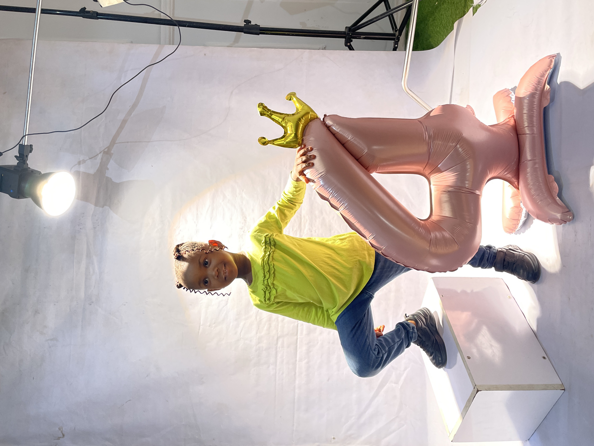 A young girl in a yellow long-sleeve shirt and jeans stands on a white box, holding a rose-gold balloon shaped like the number "4" topped with a gold crown. The setting appears to be a photography studio with a white backdrop, lights on stands, and a patch of artificial grass in the corner. The photo was taken in Jos, Nigeria.
