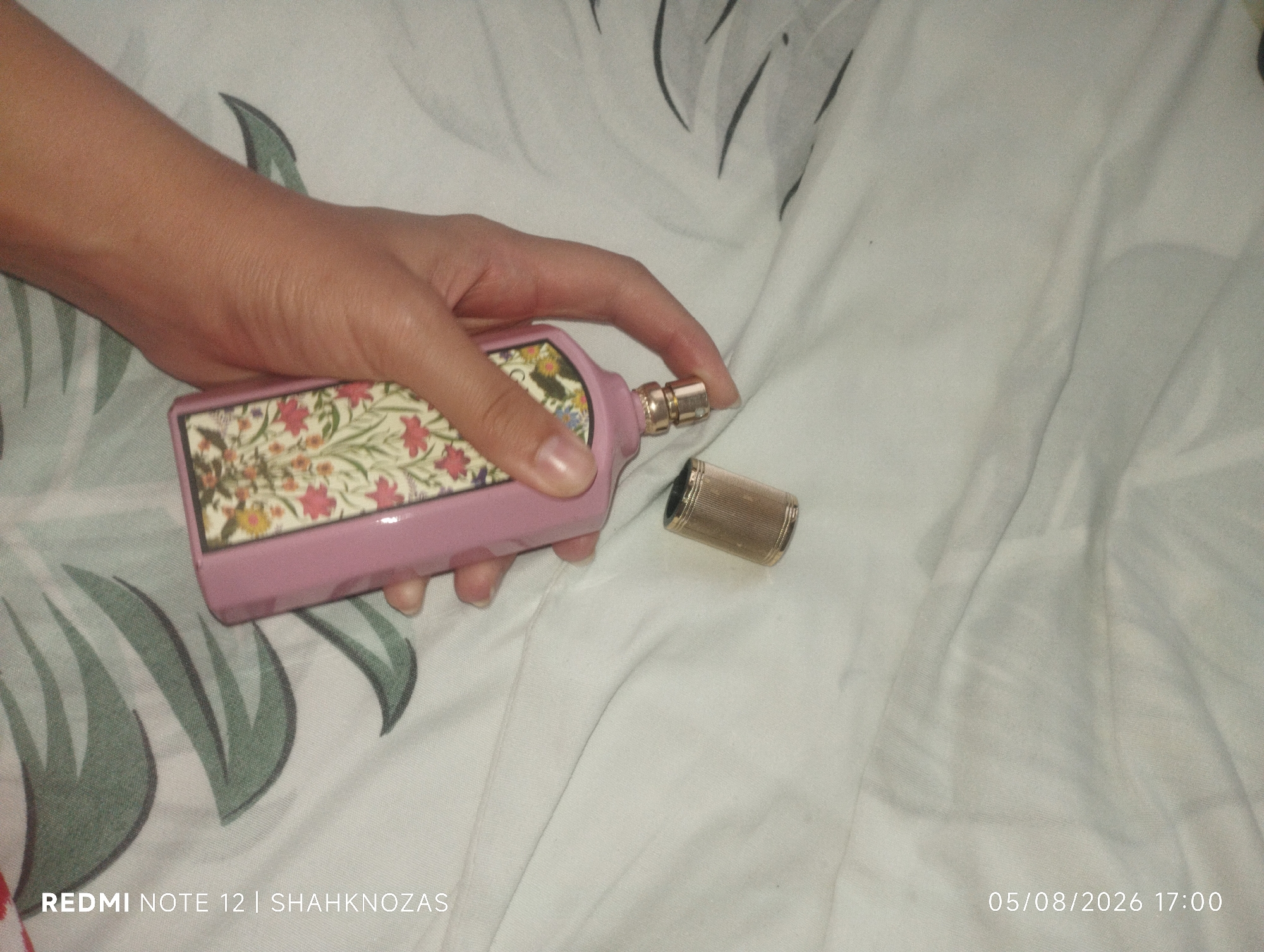 A person's hand is holding a pink perfume bottle with a floral design. The cap of the perfume bottle is off and lies next to it. The scene is set on a white, wrinkled fabric, possibly a bedsheet, with a green leaf pattern visible in the background. The timestamp "05/08/2026 17:00" and "REDMI NOTE 12 | SHAHKNOZAS" are present in the lower right and lower left corners respectively. The location is Chibintepa, Uzbekistan.