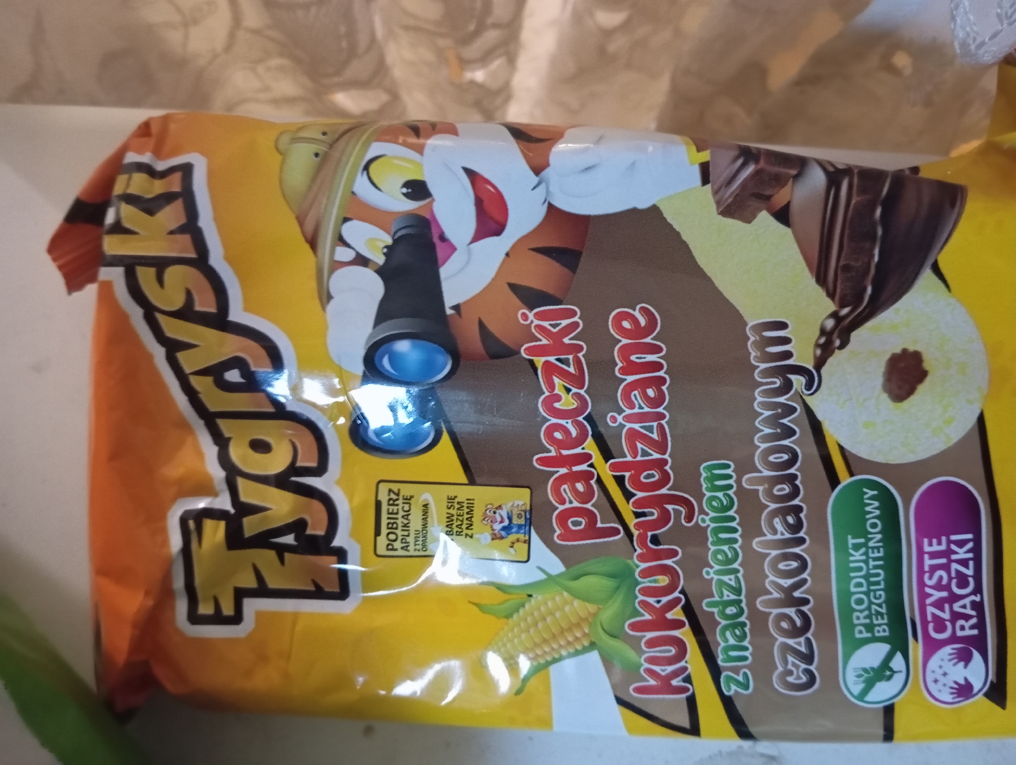 A close-up image shows the front of a yellow and orange bag of "Tygrsyk" brand corn puffs with chocolate filling. The bag features cartoon graphics of a tiger wearing a safari hat and binoculars, a corn cob, and images of the puffed snacks. Text on the bag indicates "pateczki kukurydziane z nadzieniem czekoladowym" (corn puffs with chocolate filling), "Produkt Bezglutenowy" (Gluten-Free Product), and "Czyste Raczki" (Clean Hands). The lighting is somewhat dim and casts shadows, suggesting an indoor setting. There are no people or any identifiable landmarks of Zaporizhzhia, Ukraine visible in the image.