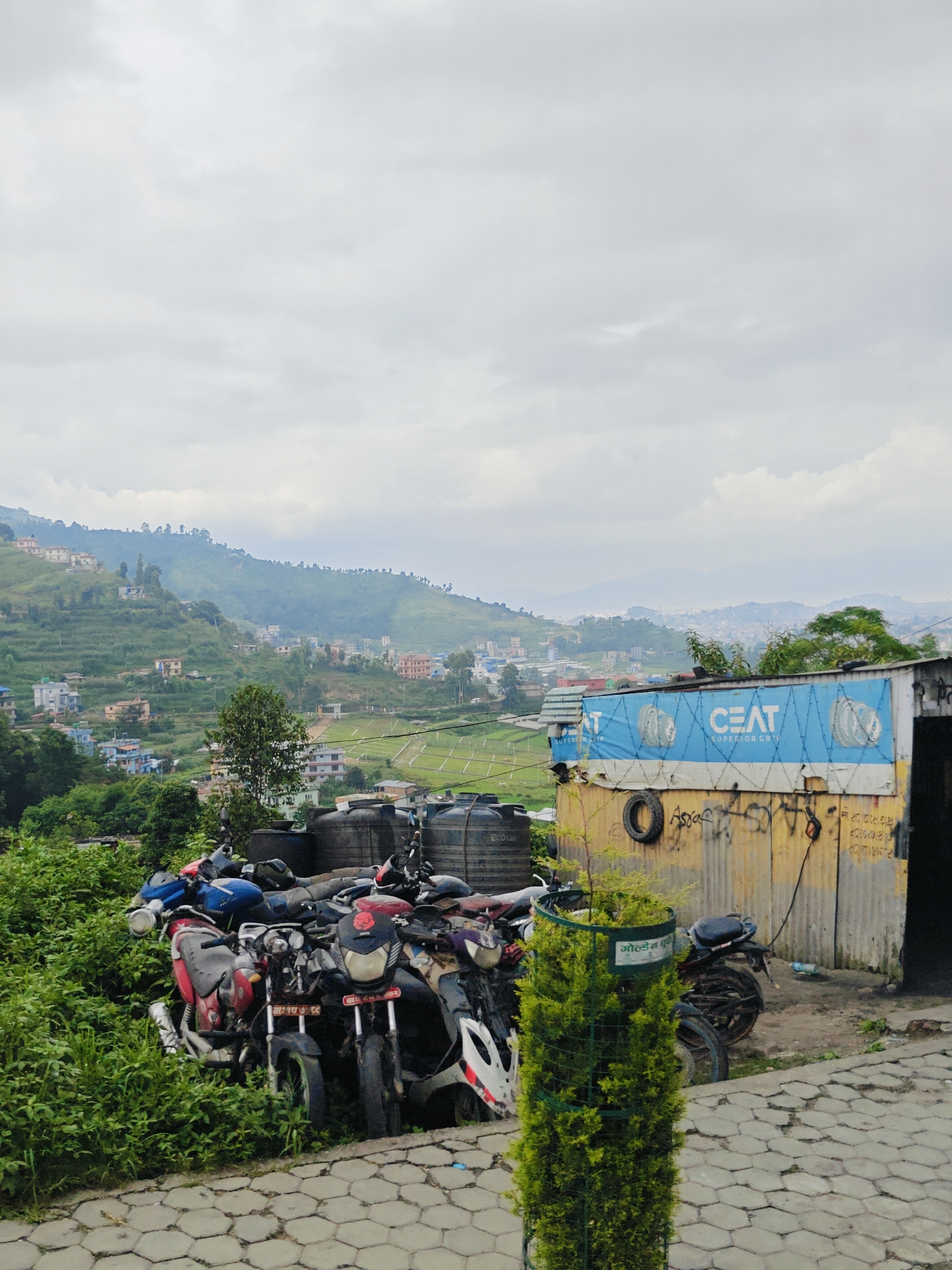 The image depicts a cloudy day in the city of Chandragiri, Nepal, with a focus on a collection of motorcycles parked in front of a corrugated metal structure. The structure has a blue tarp advertising "CEAT SUPERIOR GRIP" stretched across its front, adorned with images of tires. Below the tarp, the wall is covered in graffiti and a black spare tire is mounted on it. Several large black water tanks are stacked behind the motorcycles. The motorcycles themselves are a mix of colors including red, blue, and black, and appear to be either abandoned or awaiting repair, partially obscured by dense green foliage. In the background, rolling hills are covered with houses and terraced fields, indicating a semi-urban or rural mountainous landscape. The foreground features a paved area with hexagonal interlocking stones and a small, conical green shrub enclosed in a green metal cage with writing on it. The overall scene suggests a roadside location with a repair shop or storage area for vehicles, set against the backdrop of the mountainous terrain characteristic of Chandragiri.
