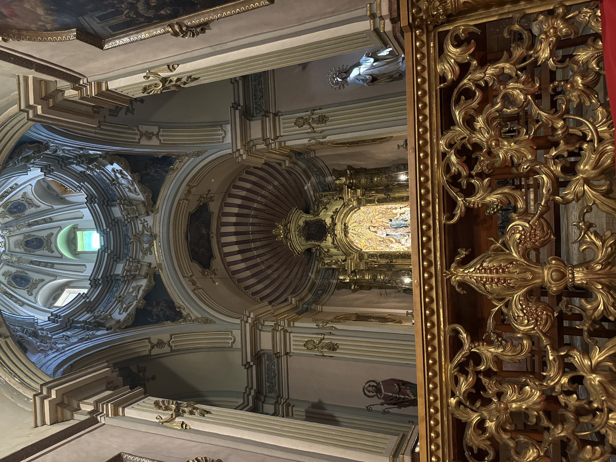 This is an interior shot of the Santuari de la Mare de Déu de la Victòria church in Sóller, Spain. The image is taken from a lower vantage point, looking up towards the altar and the church's dome. In the foreground, there is a richly ornate, gilded wooden balustrade. Behind this, a wooden railing with beaded detailing separates the nave from the sanctuary.
The sanctuary features a prominent gilded altarpiece, which is intricately carved and adorned with numerous statues. At the center of the altarpiece, a religious scene is depicted with multiple figures, including a central Madonna and child. To the left of the altarpiece, a statue of a male saint holding a staff is visible. To the right, a statue of a female saint with a halo crown is present.
The architecture of the church is grand, with towering columns, arched doorways, and decorative cornices, all highlighted with gold accents. The ceiling is a domed structure, showcasing elaborate plasterwork and painted panels. A window in the dome allows natural light to filter into the space. The walls are painted in a pale pink hue. The overall impression is one of opulence and religious devotion.