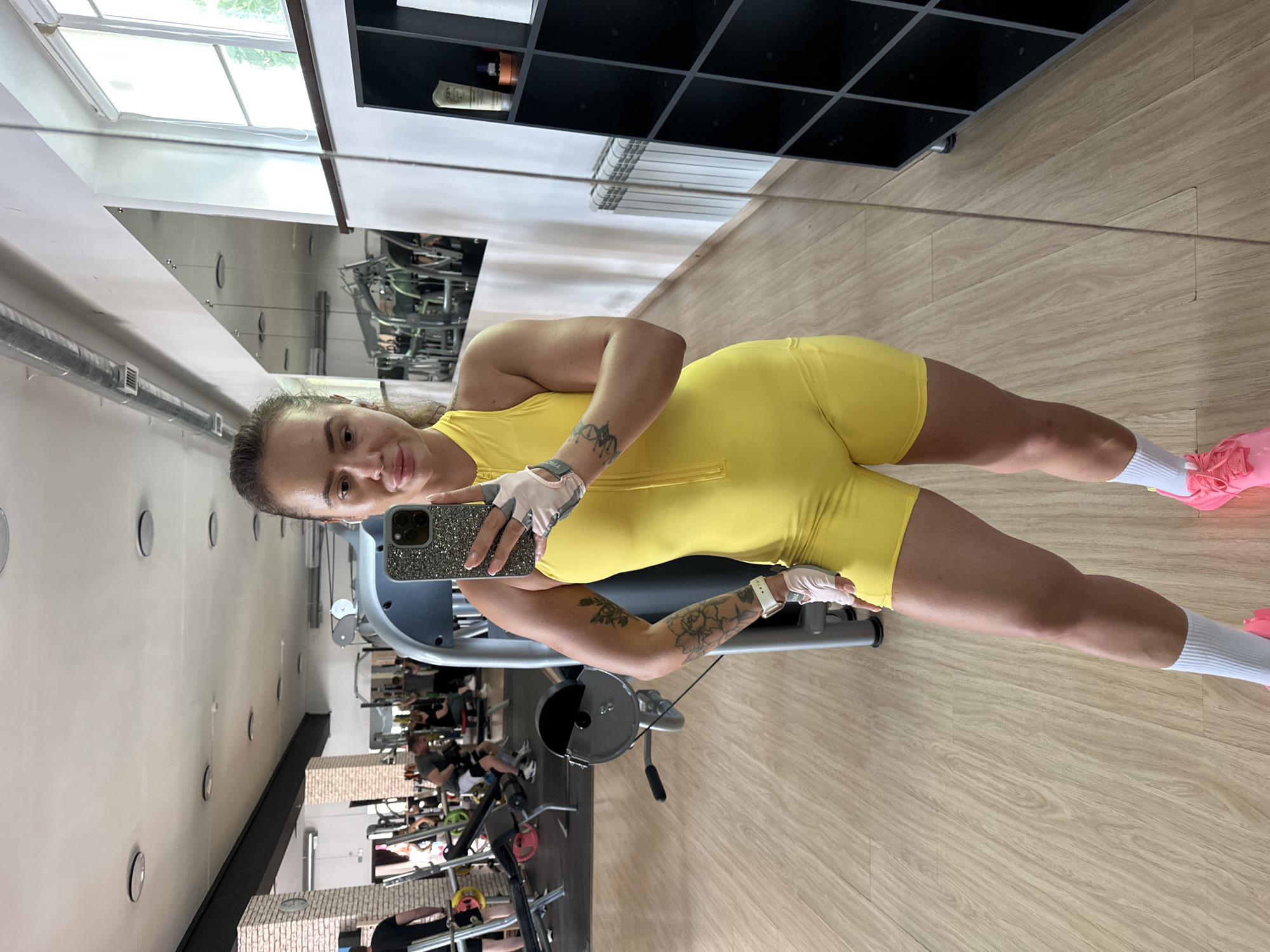 A woman is taking a mirror selfie in a gym. She is wearing a bright yellow workout outfit, white gloves, and pink athletic shoes. She has a phone in her right hand and a tattoo on her left arm. The gym has wooden floors, weight machines, and mirrors on the walls. Other people are visible in the background, exercising. The image is taken in Yuzhnouralsk, Russia.