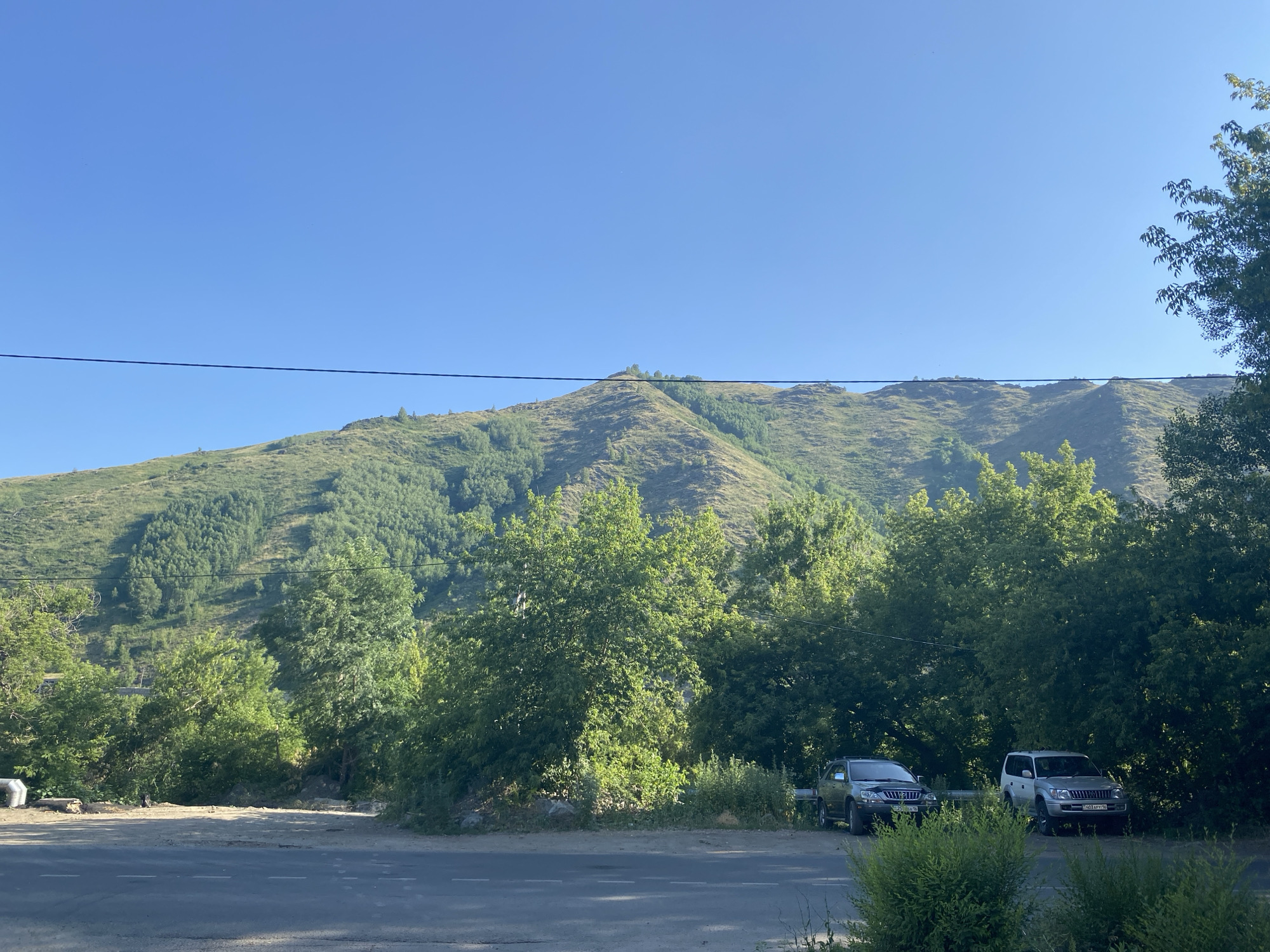 The image depicts a scenic view of a mountain range covered in lush green trees under a clear blue sky. In the foreground, there is a paved road with white dashed lines. To the right of the road, two SUVs are parked. The one on the left is a dark-colored SUV, and the one on the right is a silver SUV. Behind the vehicles, a dense line of trees partially obscures the base of the mountain. The mountain itself is steep and covered with a mixture of lighter green shrubs and darker green trees, with visible textures and contours. A thin black wire stretches horizontally across the upper portion of the frame, separating the sky from the landscape. The overall scene suggests a natural, possibly rural or mountainous area on the outskirts of a city. Given the context of Oskemen, Kazakhstan, this could be a view from a road leading into or out of the city, showcasing its proximity to natural landscapes.
