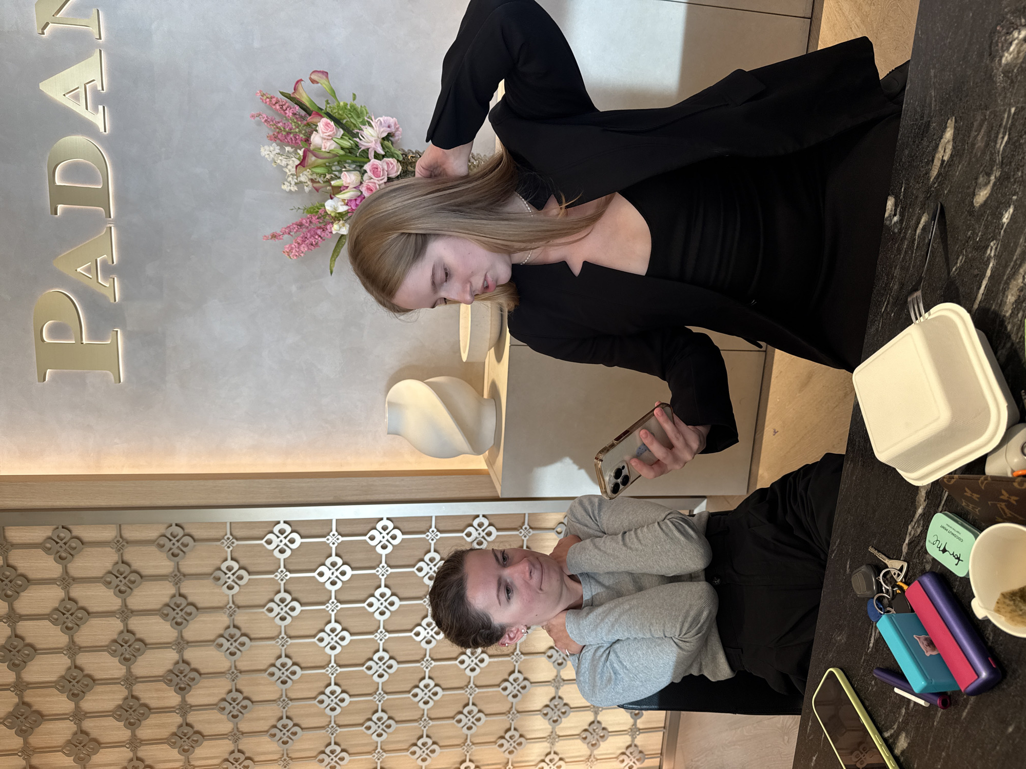 Two young women are seated at a dark marble table in what appears to be a boutique or high-end establishment, suggested by the floral arrangement and the "PADAN" signage on the wall, which is likely a brand name. The woman on the left, wearing a grey long-sleeved shirt and black pants, has her hands clasped around her neck and appears to be looking down with a concerned expression. The woman on the right, dressed in a black blazer and black dress, is holding a smartphone, seemingly showing something to the other woman, and is touching her hair. On the table, a variety of objects are present: a yellow-cased smartphone, a blue and pink electronic device, a set of keys, a small mint tin, a white takeout container with a fork, a brown patterned item, and a white teacup with a tea bag. The background features decorative wooden paneling on the left and a textured grey wall with lighting and a sculptural vase on a shelf to the right. The scene suggests a moment of shared attention, possibly concern or surprise, between the two individuals. There is no explicit visual information to confirm the specific location within Tel-Aviv, Israel, beyond the possibility that "PADAN" might be a local business.