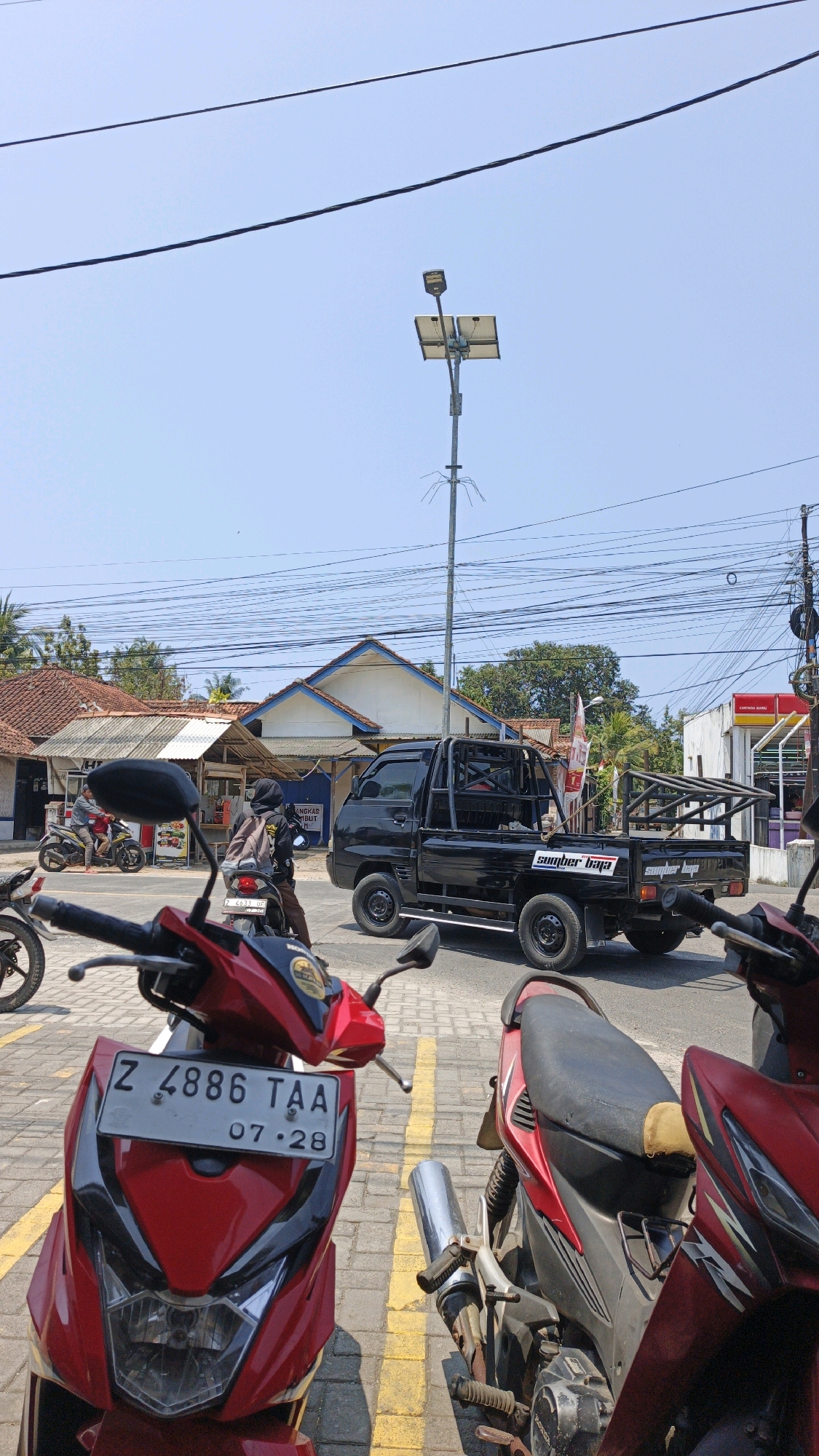 The image captures a street scene in Cibenda, Indonesia, under a clear blue sky. In the foreground, the front of two red motorcycles is visible, one with a license plate reading "Z 4886 TAA 07-28". Behind them, a black pickup truck with a reinforced bed is parked on the street, with "sumber baja" written on its side. Further in the background, a solar-powered street lamp stands tall with its panels angled towards the sun. Several buildings with terracotta roofs are visible, some appearing to be shops or homes. Power lines crisscross the sky above the street. A few individuals are present, one riding a motorcycle and another walking near the shops. The ground is paved with interlocking bricks. The overall impression is of a typical day in a semi-urban Indonesian setting.