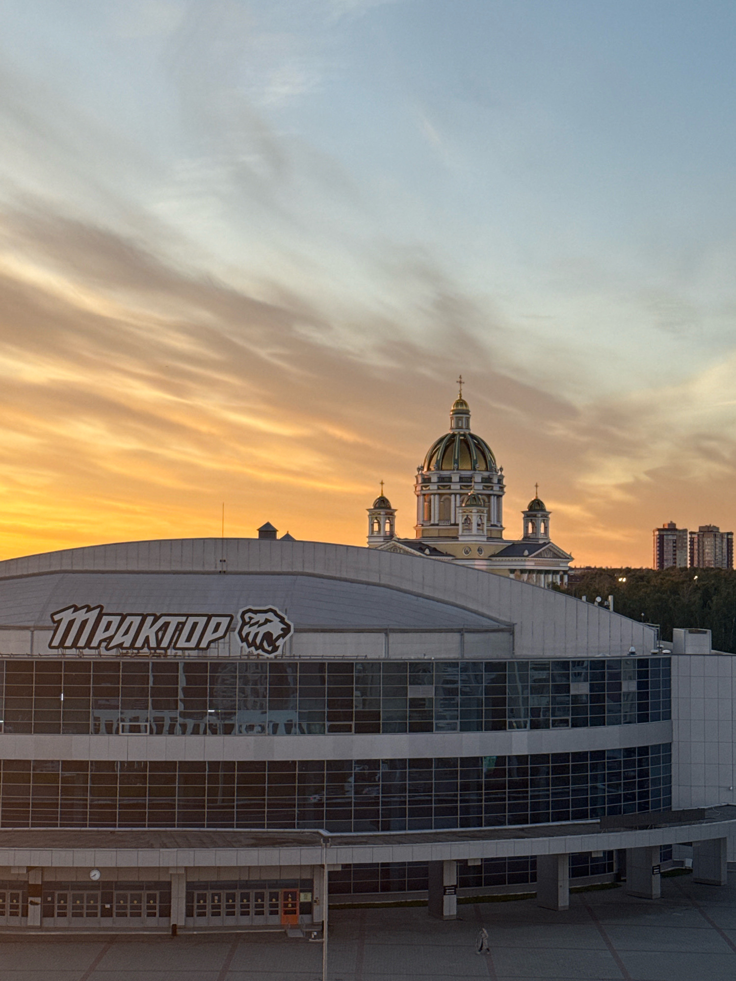 The image shows a wide shot of the Traktor Ice Arena in Chelyabinsk, Russia, during a sunset. The sky is filled with dramatic orange and yellow clouds, transitioning to a pale blue at the top. In the background, a prominent Orthodox church with golden domes and crosses stands out against the sky. To the right of the church, modern residential buildings can be seen.
The Traktor Ice Arena, the main subject, is a large, modern building with a distinctive rounded roof. Its facade is primarily glass, reflecting the sky and the surrounding structures. A prominent sign on the upper part of the building reads "ТРАКТОР" in bold white letters, accompanied by a stylized image of a wildcat, the symbol of the Traktor Chelyabinsk ice hockey team.
Below the main arena structure, there is an entrance area with multiple doors. A clock is visible above one of the entrances. In the foreground, a paved plaza is visible, and a single person in light-colored clothing is walking in the distance. The overall impression is of a city scene at dusk, with a blend of architectural styles and a dramatic natural backdrop.
