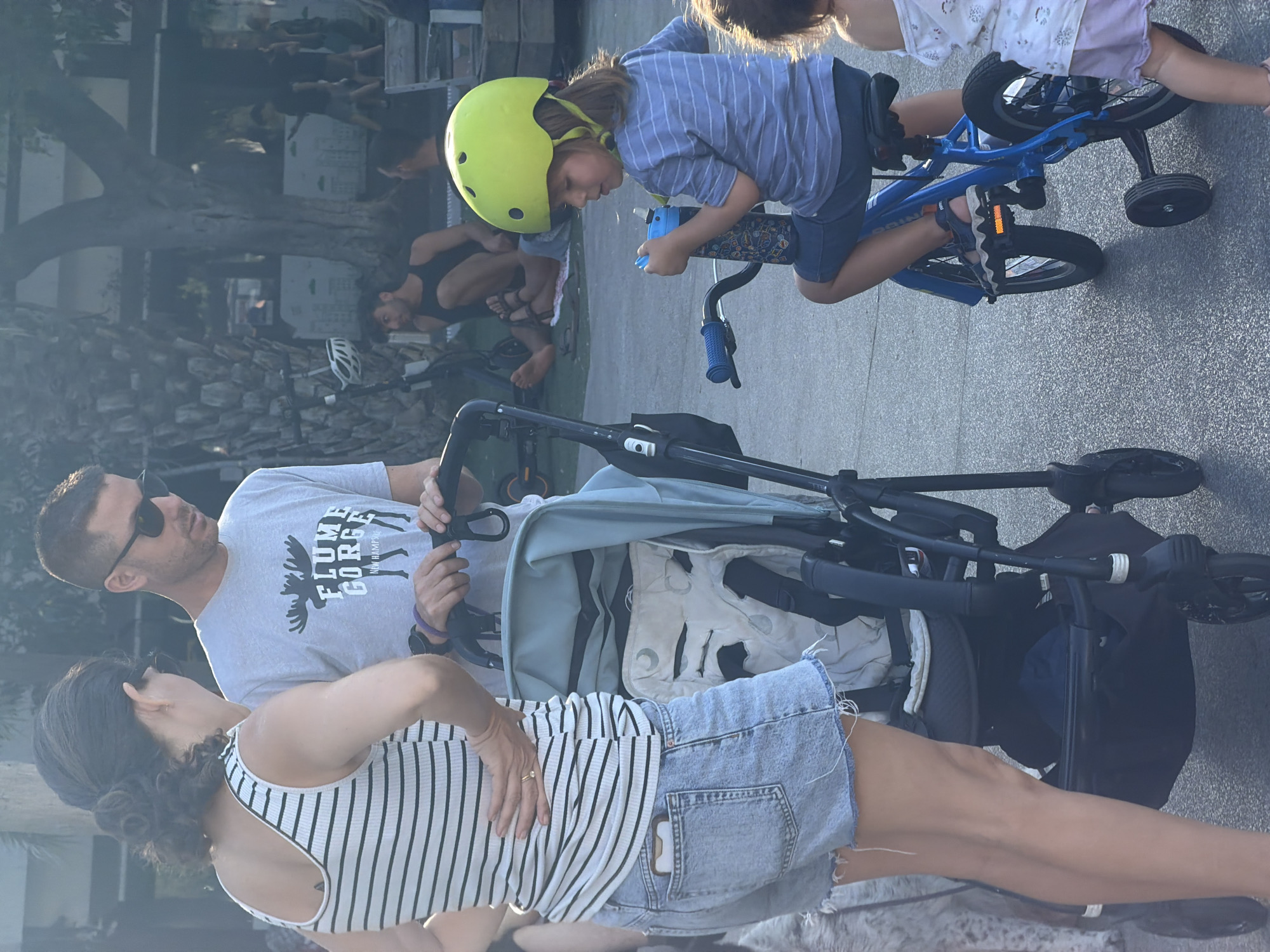 This outdoor daytime image depicts a family of four, including two adults and two children, in a public area of Tel-Aviv, Israel. A woman stands on the left, facing right, with her right hand on her hip, looking towards the man. She has dark hair pulled back, wears a white and black striped sleeveless top, light blue denim shorts with cut-off hems, and a gold ring on her left ring finger; a smartphone is visible in her right rear shorts pocket. To her right, a man stands holding the handle of a black baby stroller with a light blue canopy and a patterned seat insert. He has short dark hair, stubble, wears dark sunglasses, a light gray t-shirt with a dark "FLUME GORGE NEW HAMPSHIRE" moose graphic, and a dark wristwatch on his left wrist; he is looking down. To the right of the stroller, a young child, approximately 4-6 years old, sits on a blue bicycle with training wheels, wearing a bright neon yellow helmet, a blue and white striped t-shirt, dark shorts, and sandals; the child is holding a dark blue water bottle to their mouth. Partially visible on the far right is a second, younger child wearing a white top and light purple bottoms. In the background, several individuals are visible; one man in a black tank top is seated on the ground, barefoot, near a large palm tree. Other trees and a paved surface are present, illuminated by bright daylight.