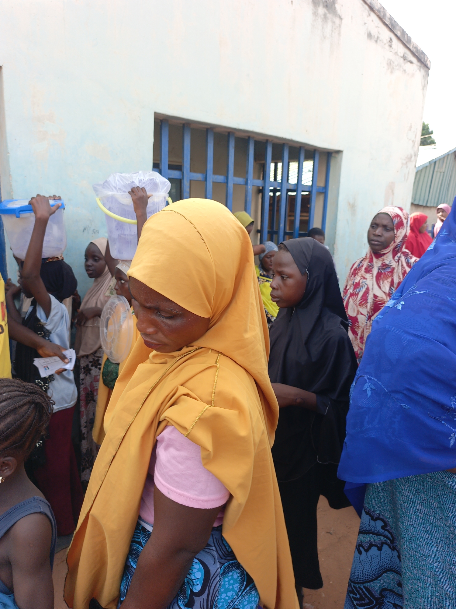 The image shows a group of people in Lafia, Nigeria. Most of the people are women and girls, and many are wearing hijabs or other traditional Islamic clothing. Some are carrying buckets. They are standing outside a light blue building with a barred window. The environment seems to be a street or public area.