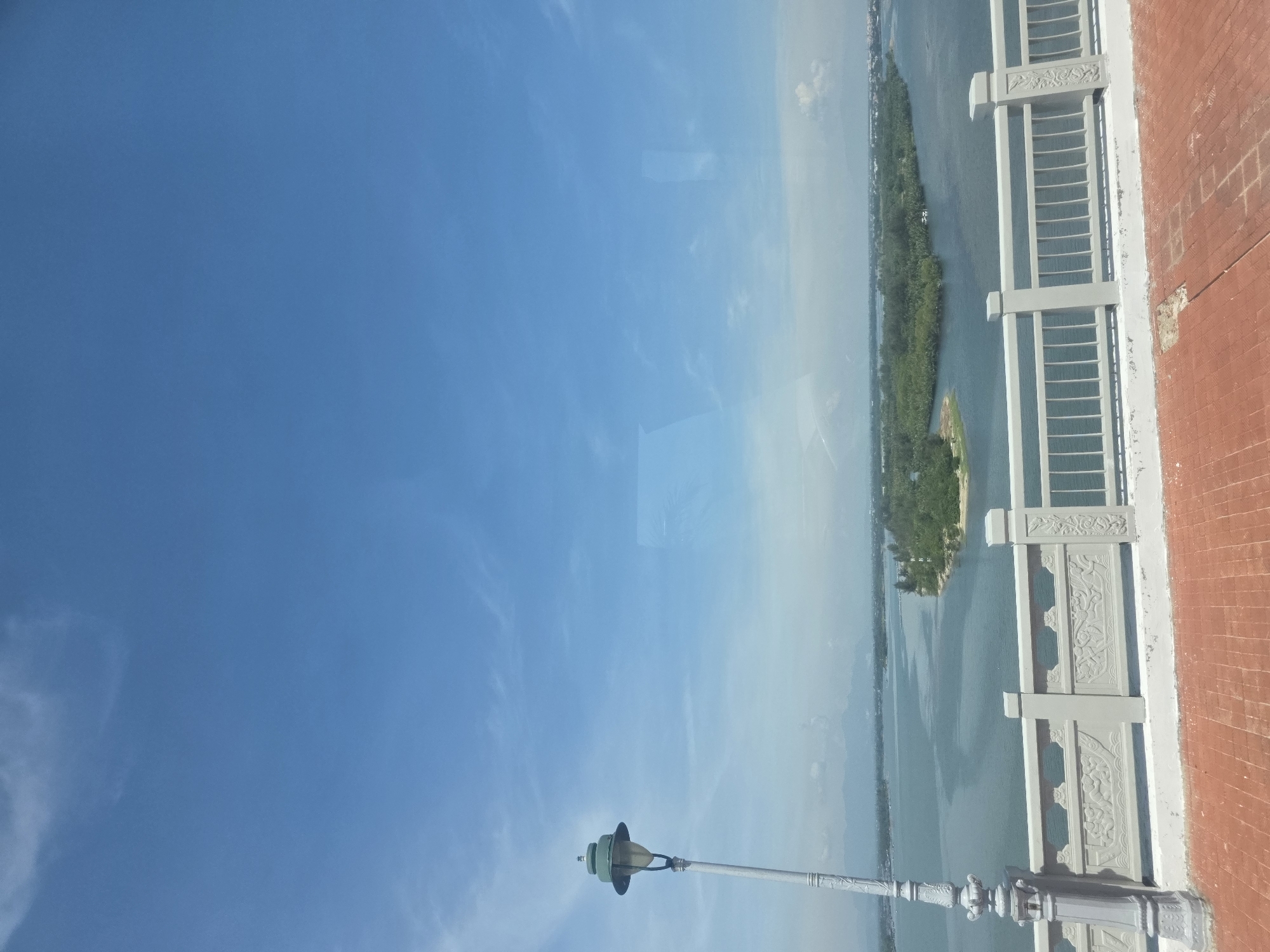 The image shows a view from a high vantage point, possibly a building or a bridge, overlooking a body of water with a narrow, tree-covered island in the middle. The sky is blue with some light clouds. In the foreground, there is a decorative concrete railing and a section of a red brick wall. A street lamp stands in the foreground along the railing. It is likely the Hàn River and possibly the view from the Dragon Bridge in Đà Nẵng, Vietnam.