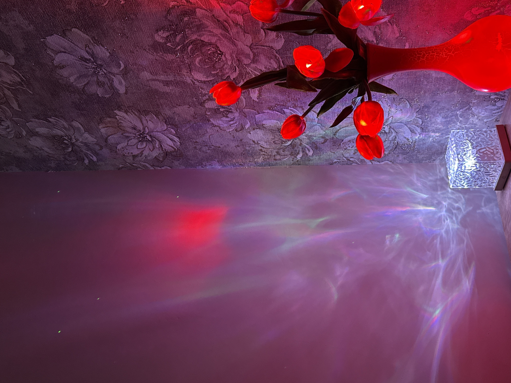The image shows a room with floral wallpaper on the upper half and a plain wall on the lower half. On the right side of the floral wallpaper, there's a red vase with red tulip-like flowers. Below the vase, there's a decorative box with geometric cut-outs. The light from the box and flowers is projected onto the plain wall, creating a rainbow-like effect. There are no people in the image. The location of the city is Unknown, Unknown.