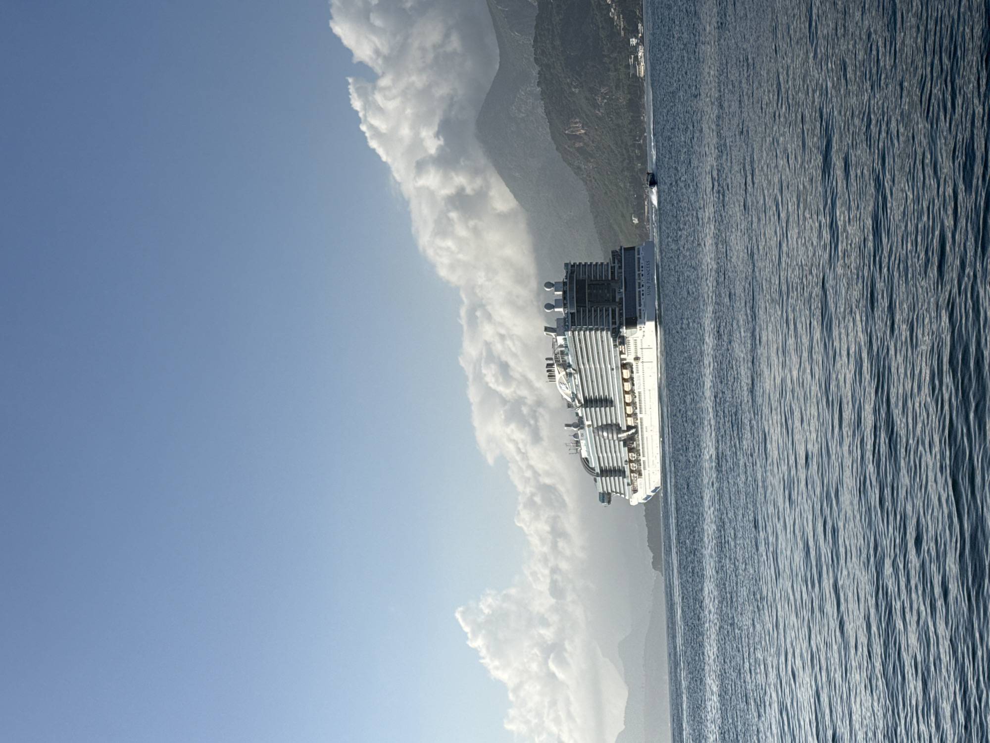 The image depicts a large cruise ship, identified by its hull name "Sun Princess," sailing along the coast of Bar, Montenegro. The ship is seen from a side perspective, suggesting it's either passing by or approaching the coastal area. The topography indicates mountainous terrain along the coastline, partially obscured by cloud cover. A small village or town is visible along the coast, further confirming the location. The sea stretches to the right of the ship, with visible water texture. The sky occupies the upper portion of the image, with a clear blue expanse above the coastal clouds.