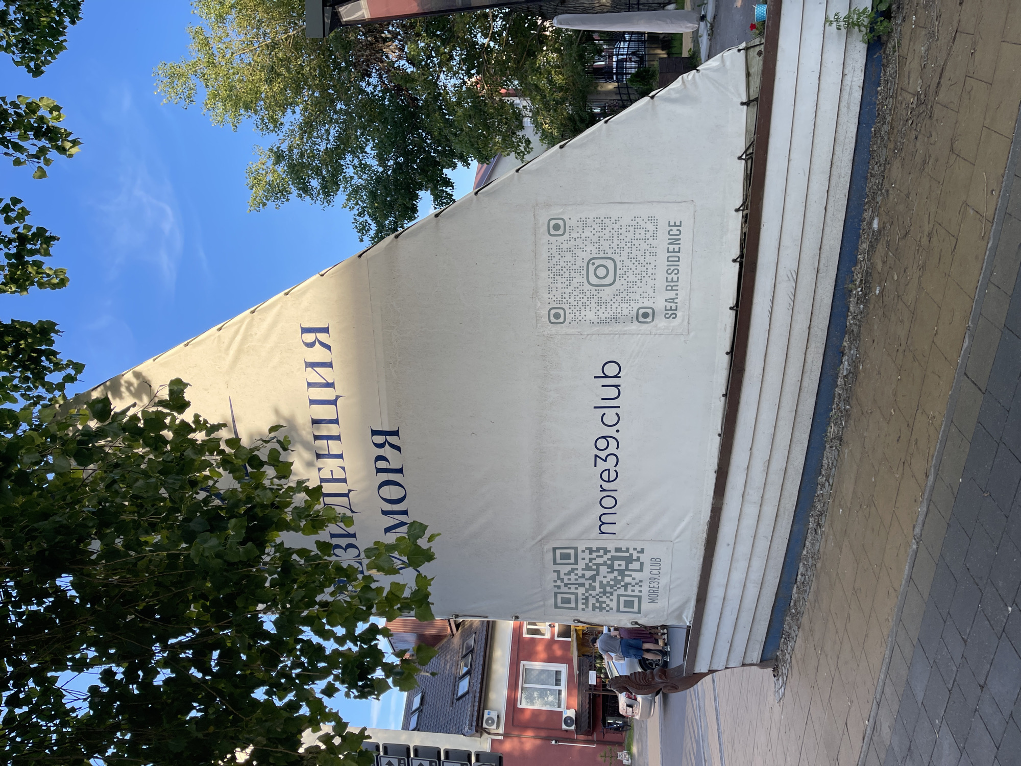 In Zelenogradsk, Russia, a white sail on a boat-shaped structure displays blue Cyrillic text reading "РЕЗИДЕНЦИЯ МОРЯ" (Residency of the Sea). Two QR codes are visible: one on the left with the text "MORE39.CLUB" beneath it, and another on the right with an Instagram logo and "SEA.RESIDENCE" below. A person in a red building with visible air conditioning units stands near the left QR code. A car is parked on the street. Trees with green leaves overhang the scene, and the sky is blue.