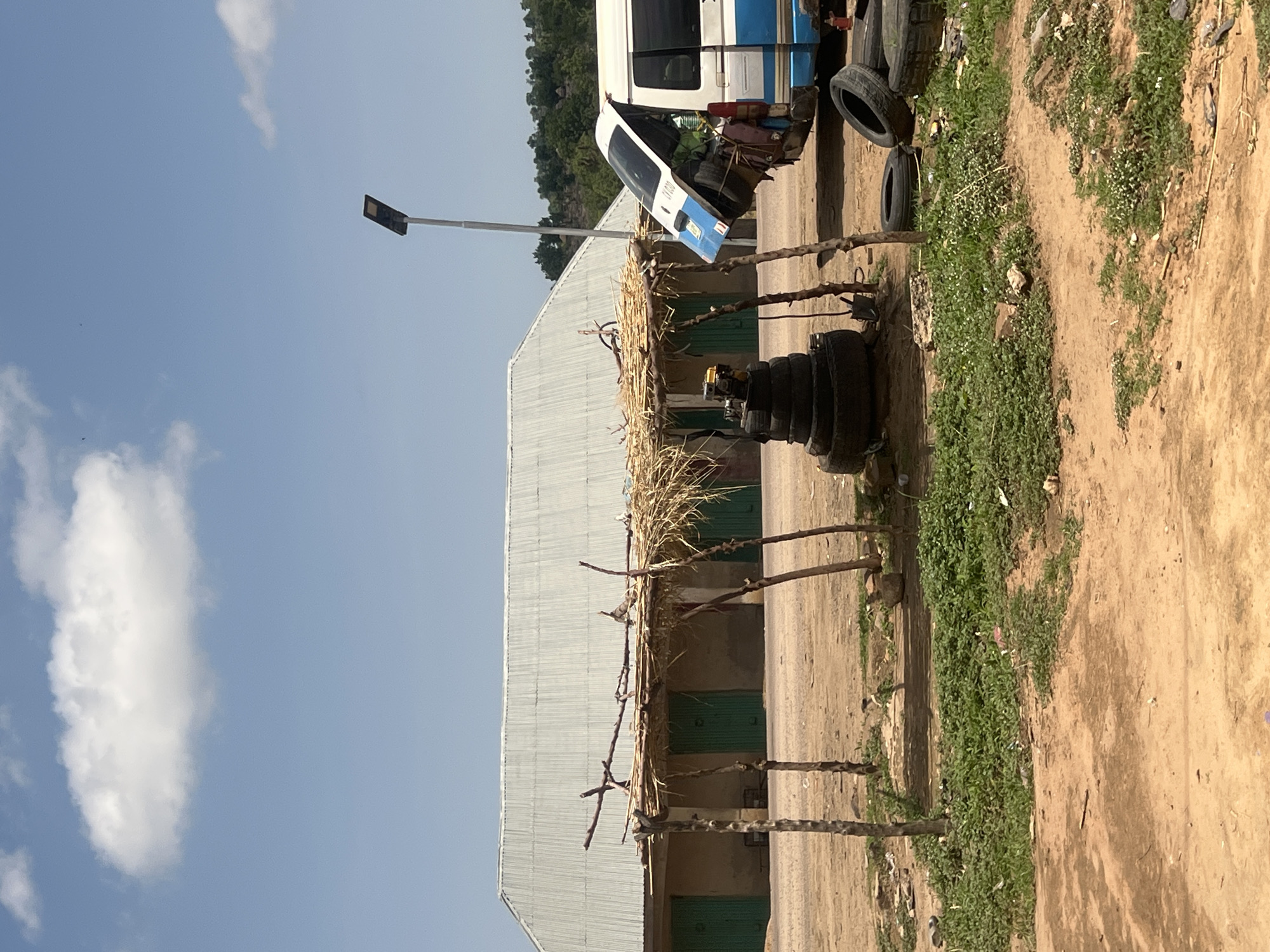 The image depicts an outdoor scene in Sabon Layi Jessu, Nigeria, under a clear, bright blue sky with scattered white cumulus clouds. In the foreground, the ground is composed of reddish-brown dirt and sand, with patches of green, low-lying vegetation.
A makeshift, open-sided shelter stands centrally, constructed from rough wooden poles supporting a sparse thatched roof made of dried straw or grass. Beneath this shelter, a stack of approximately six black vehicle tires forms a conical pile, topped by a small yellow and black mechanical device, possibly an engine or generator. A dark-bladed tool, resembling a shovel, is partially visible stuck into the ground near the tire stack.
Behind the shelter, a long, single-story building with light-colored walls and a corrugated metal roof is visible. The building features several identical green, double-leaf doors along its front facade.
To the right of the shelter, a white and blue minibus or passenger van is parked. The rear hatch of the van is open and tilted upwards, revealing contents packed within. The vehicle's front passenger-side door also appears to be open or removed, exposing interior components. A partially legible blue and white marker or license plate is affixed to the vehicle's rear. Several loose black vehicle tires are scattered on the ground near the van, along with a red cylindrical object.
A tall, slender pole with a dark, flat, solar-powered LED streetlight fixture on top stands between the shelter and the van. In the far background, behind the building and the van, a range of green, tree-covered hills is visible. No individuals are present in the scene.
