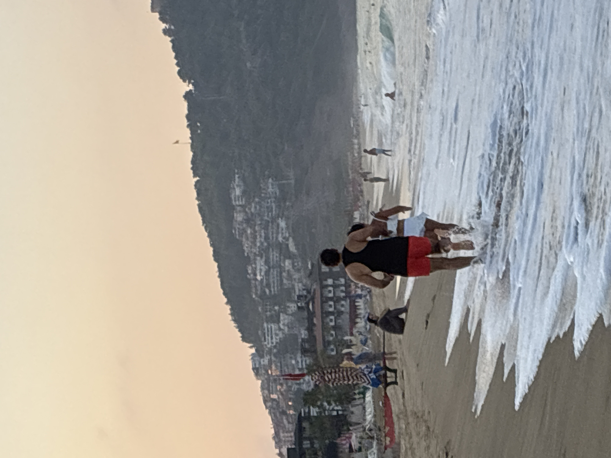 The image depicts a wide shot of a beach scene during what appears to be sunset in Alanya, Turkey. In the foreground, a man in a black tank top and red shorts and a woman in a white outfit are running into the sea. The water is shallow and foamy, with small waves lapping at the shore. In the mid-ground, other beachgoers are visible; some are standing in the water, while others are sitting on the sand. Beach umbrellas and lounge chairs are set up further back on the sand, suggesting a populated recreational area. Behind the beach, a hillside is densely covered with buildings, characteristic of the urban development in Alanya. A flag is visible on the hillside, and a castle or fortification can be seen perched at the top of the hill in the distance. The sky is a soft, hazy orange, indicative of the evening twilight.