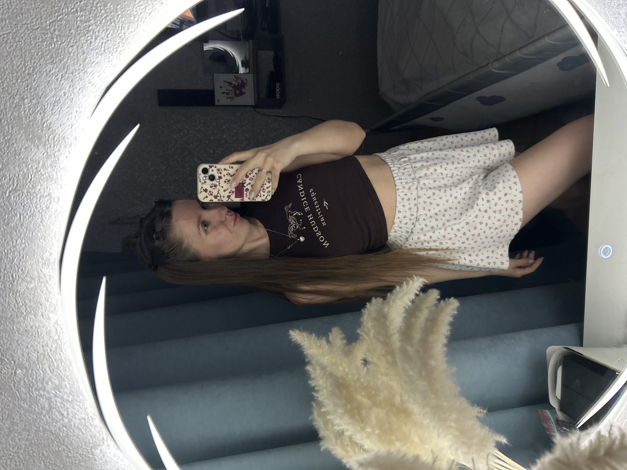 A mirror selfie captures a young woman looking into her smartphone. The woman, with long, straight brown hair, is positioned in the upper left of the circular mirror's reflection. She is wearing a dark brown crop top with reversed white text and an illustration of a stylized animal, possibly a deer or griffin. The reversed text reads "SNOGNUD" above the illustration and "CANDICE HUDSON" below it. She also wears a necklace with a pendant, rings on her fingers, and has painted nails. Her phone, held in her right hand, has a floral-patterned case.
Her attire also includes a white skirt adorned with a small floral pattern. The background reflected in the mirror shows a room with various details. To the upper right, part of a bed with a patterned mattress cover is visible. Below the bed, there is a dark piece of furniture, possibly a shelf, holding a few items including what appears to be a small lamp and a framed piece of art or card with handprints, next to a black rectangular device with "0000" and other indiscernible text.
In the foreground of the reflection, a vase filled with dried pampas grass stands to the lower right. The mirror itself is circular, featuring integrated LED lights that form elegant, curved patterns around its perimeter. The wall behind the mirror has a textured, silvery, and somewhat bumpy surface. The overall setting suggests an indoor, personal space, likely a bedroom. The LED lights on the mirror are illuminated.