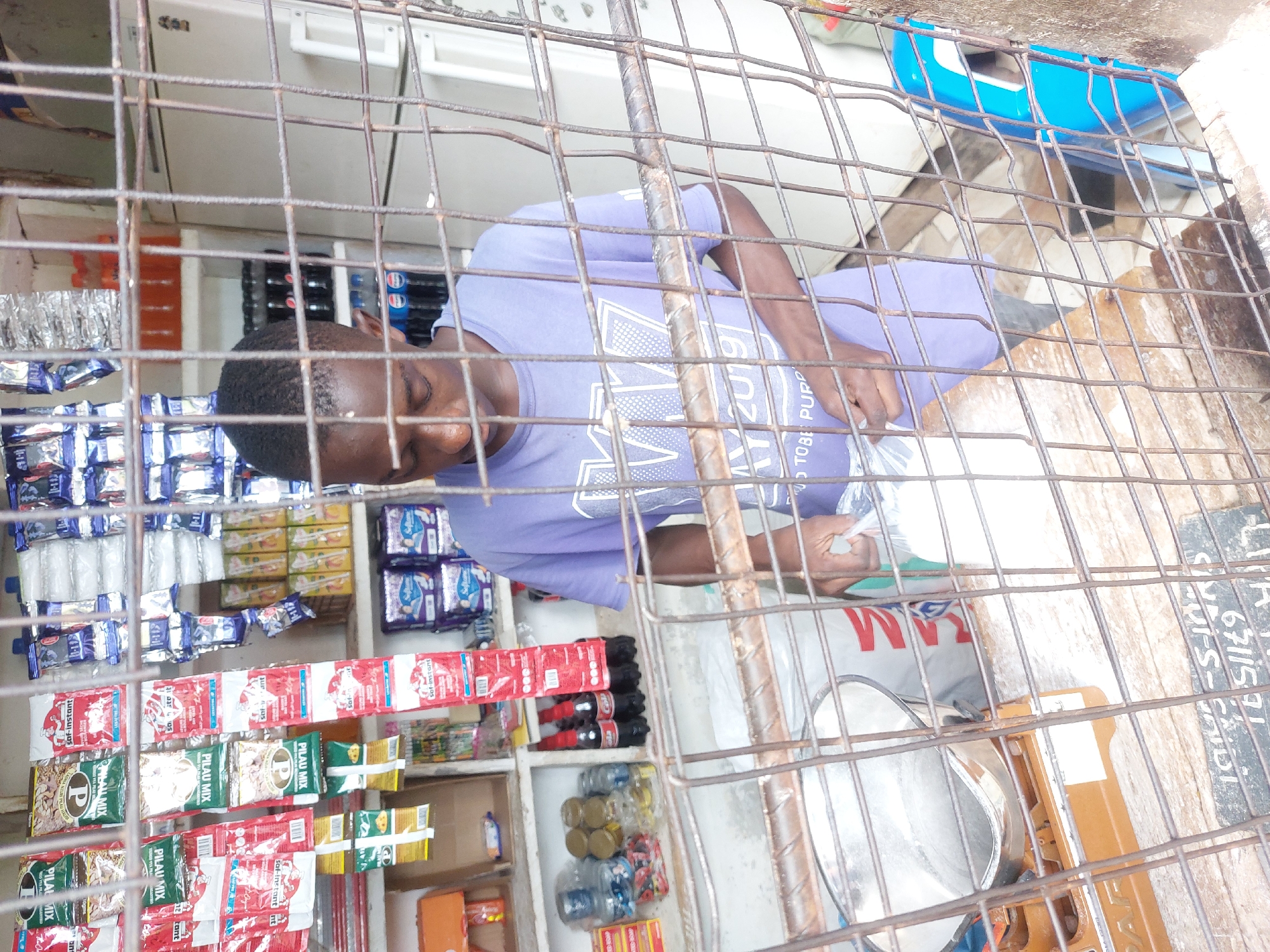 This image depicts the interior of a small retail shop or stall, likely located in Dar es-Salaam, Tanzania, as viewed through a protective metal security grid. The scene is brightly lit, suggesting daytime.
The main subject is a young man with short, dark hair, wearing a light purple t-shirt. He is standing behind a counter, looking down at an item he holds in his hands, possibly a mobile phone or merchandise. The text "MAY 2019" and "TOBE PUPP" are visible on his t-shirt, along with "AM".
The shop is stocked with various goods displayed on shelves. Prominently featured are multiple sachets of "saf-instant" and "PILAU MIX" (some with just a "P" logo), suggesting common food items. Bottled beverages, including Pepsi Cola and other sodas, along with water bottles, are also visible on shelves and in a white refrigerator/cooler in the background. Other small packaged items in foil and plastic wrappers fill the shelves.
On the counter to the man's right, a metallic weighing scale with an orange base is visible, indicating that some goods might be sold by weight. In the background, beyond the counter, a blue plastic basin or container can be seen. On a white surface to the far right, the numbers "67115" are visible. The metal grid in the foreground acts as a barrier, a common feature for security or transaction purposes in such establishments.